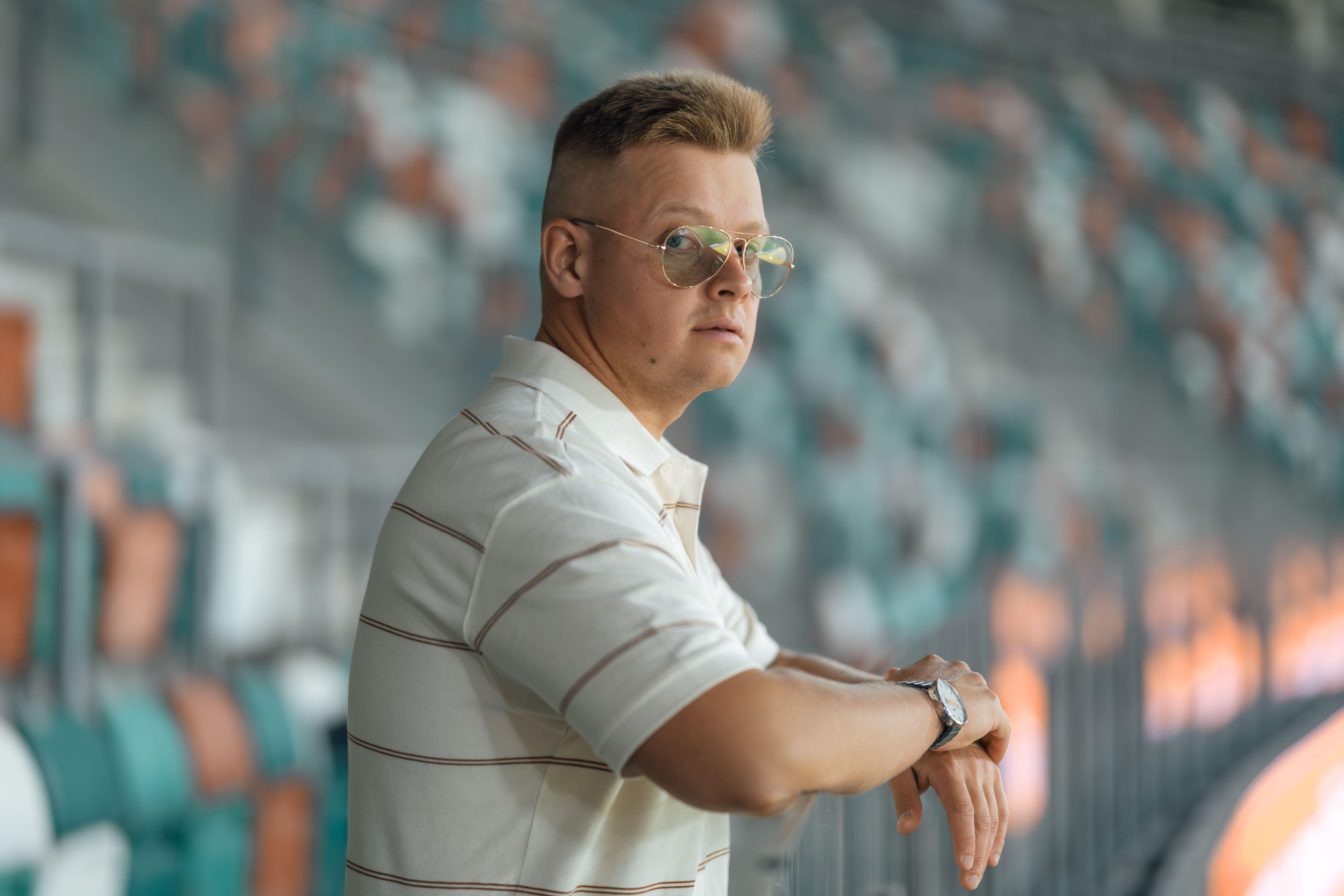 Man in white shirt wearing sunglasses, standing at an outdoor stadium with blurred seating in the background.