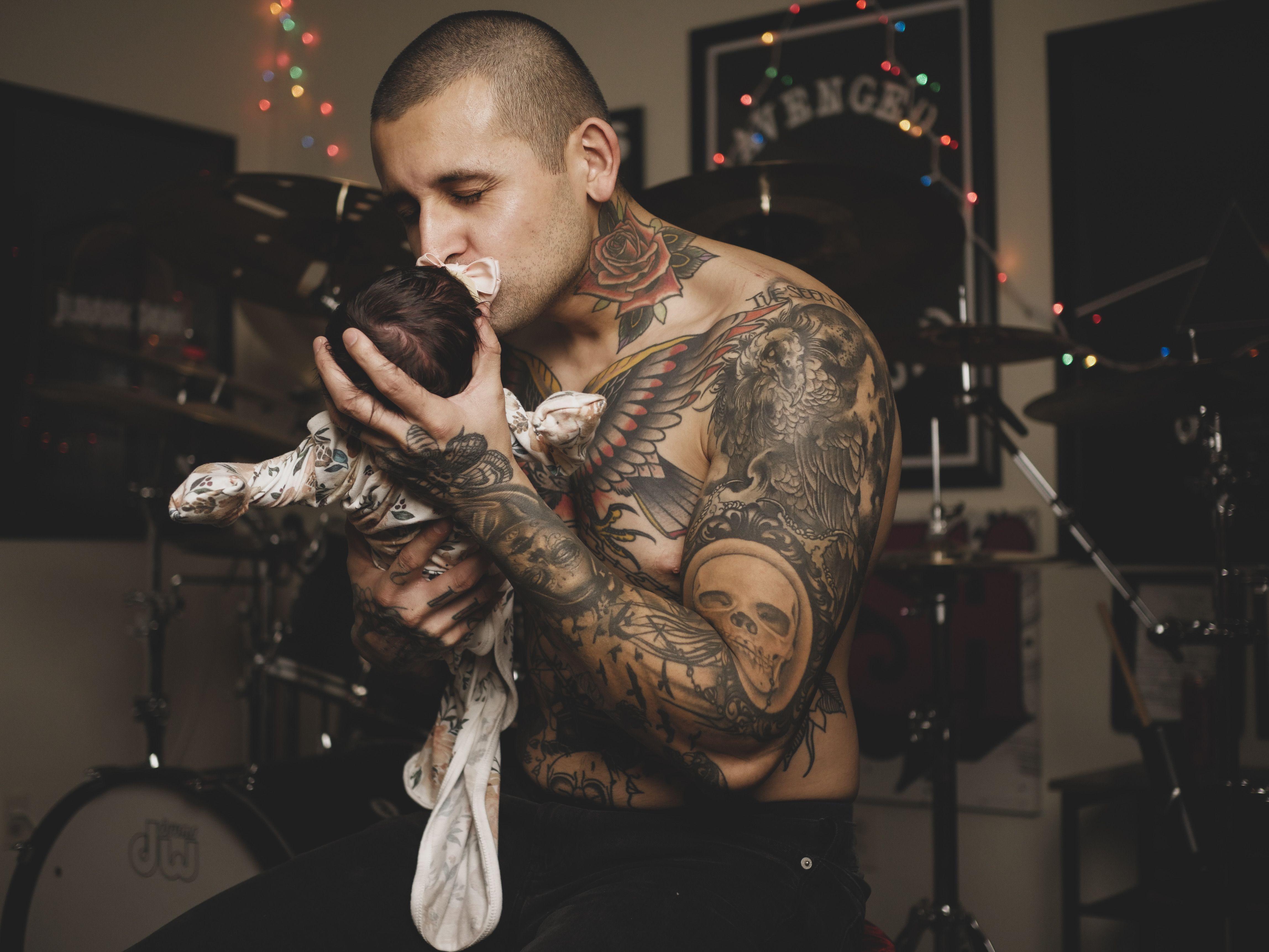 Tattooed father lovingly kisses newborn baby in a cozy room with musical instruments.