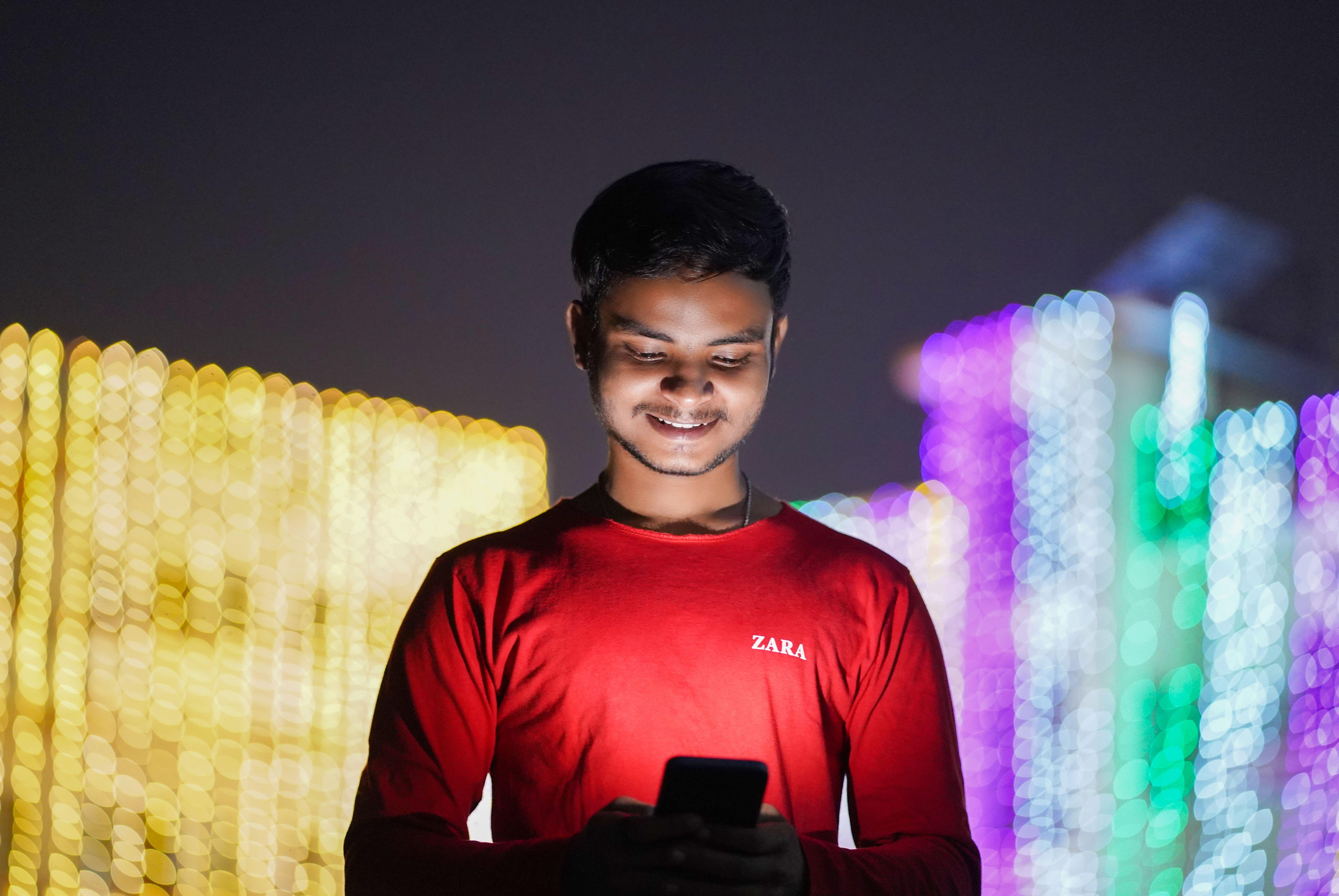 Smiling young man in red shirt using smartphone against a backdrop of festive lights at night.