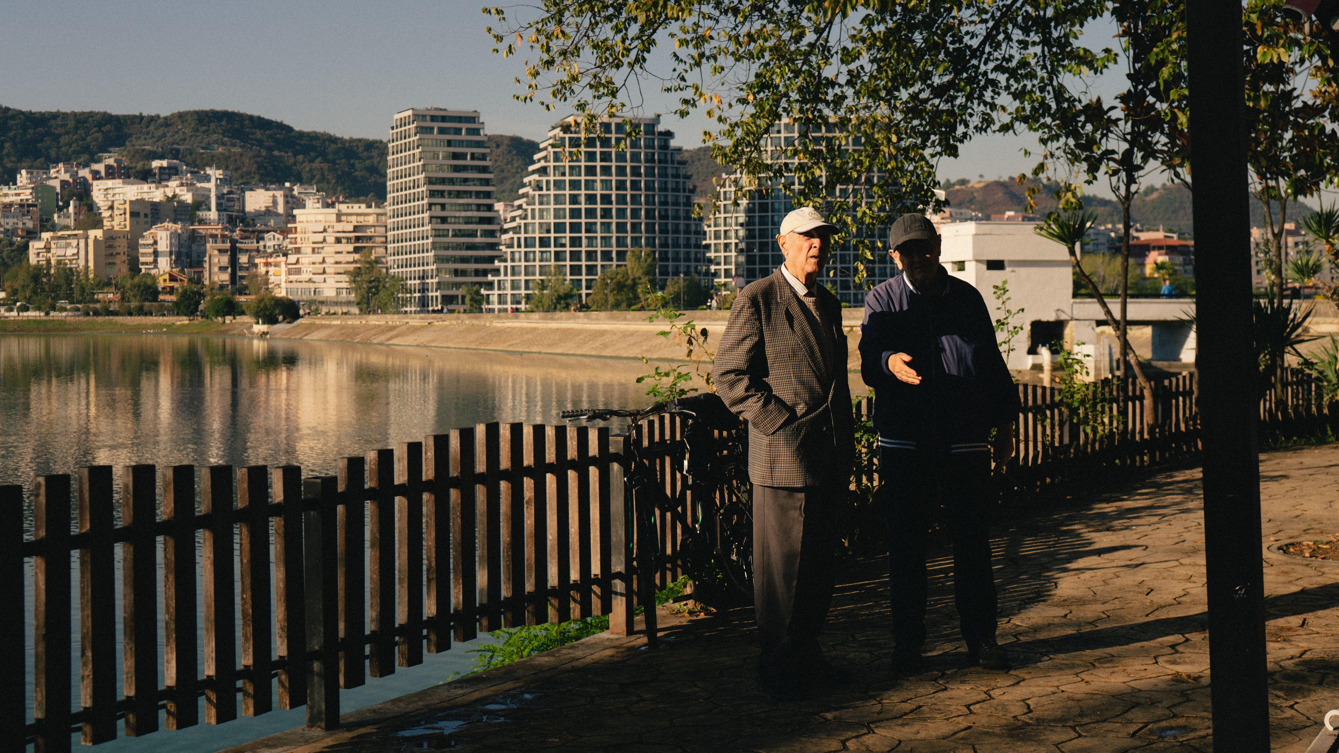 Two senior men engage in conversation along the serene Tirana lakefront, shadowed by modern architecture.
