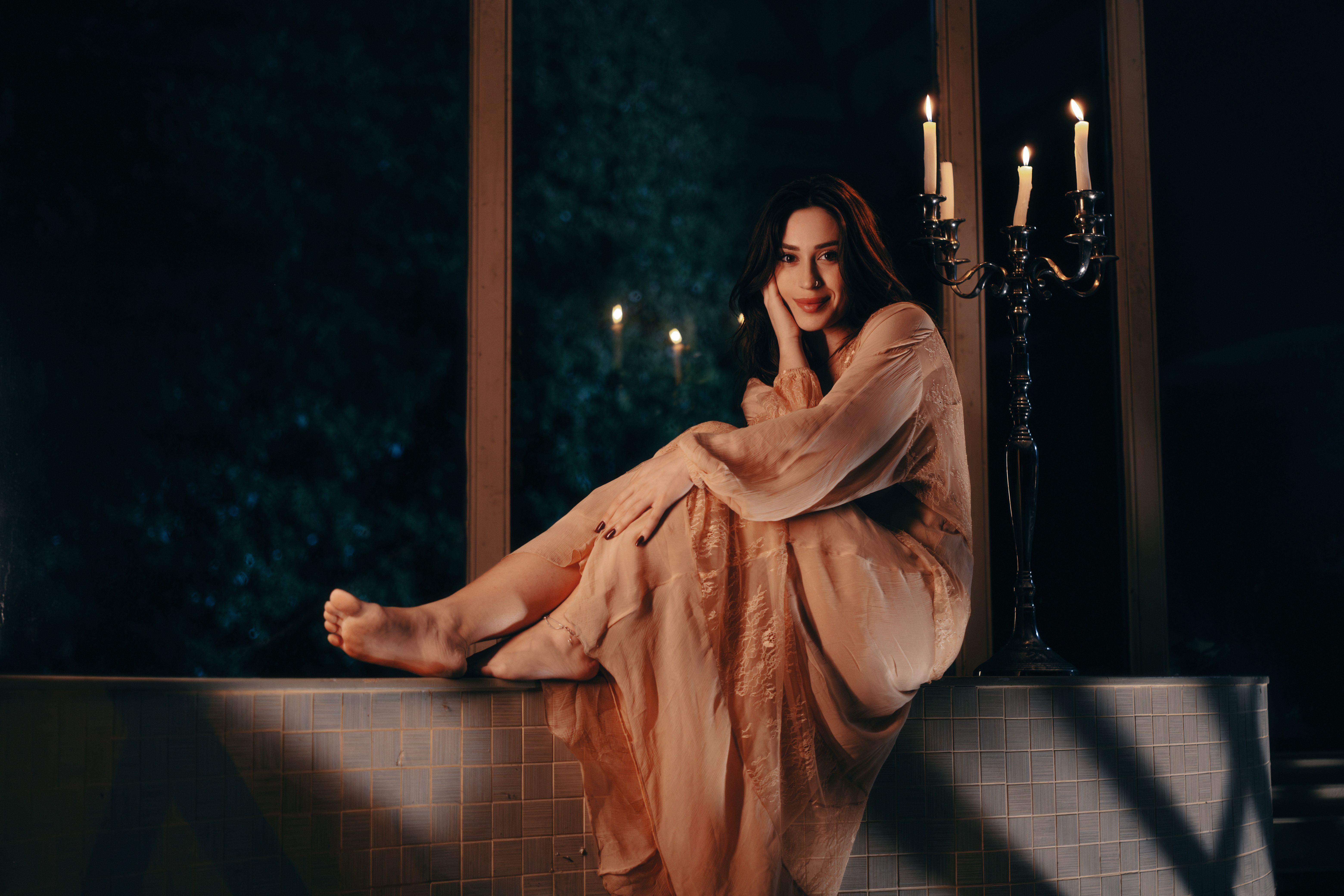 A serene portrait of a woman in a nightgown, seated beside candles, exuding elegance and calm.