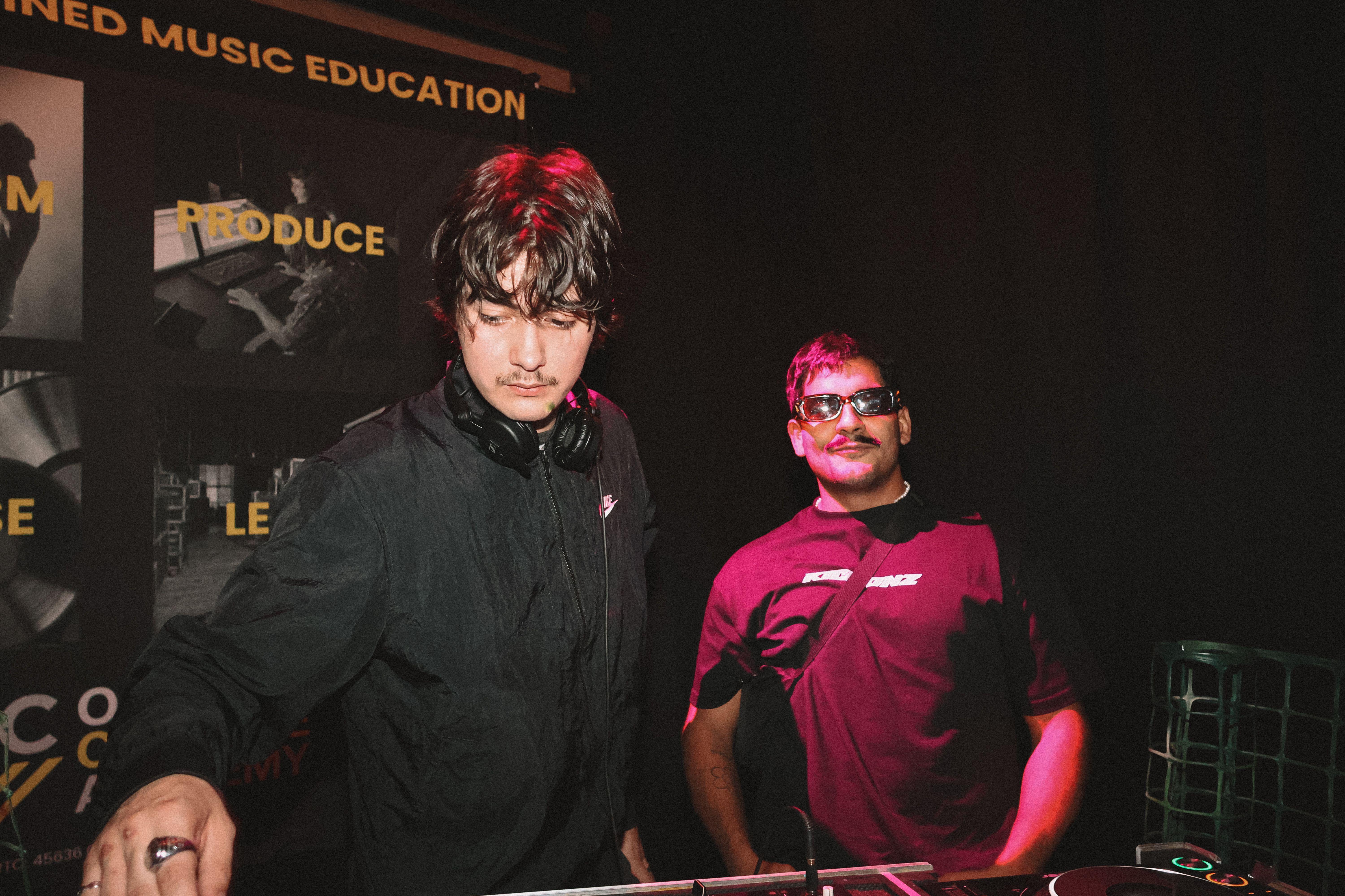 Two DJs performing at a night event with professional audio equipment and music education banner.