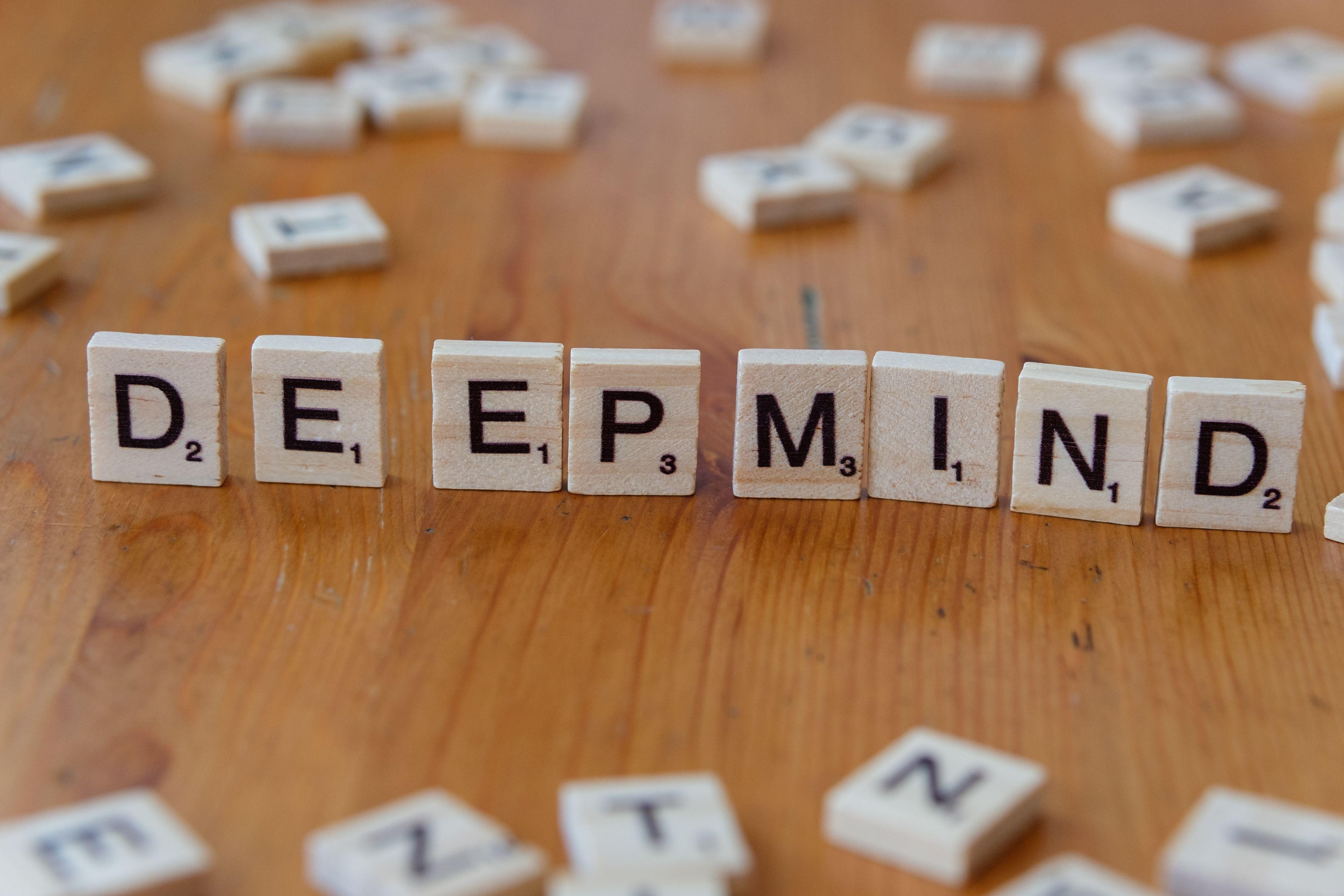 Close-up of Scrabble tiles spelling 'DeepMind' arranged on a wooden surface, with scattered tiles in the background.