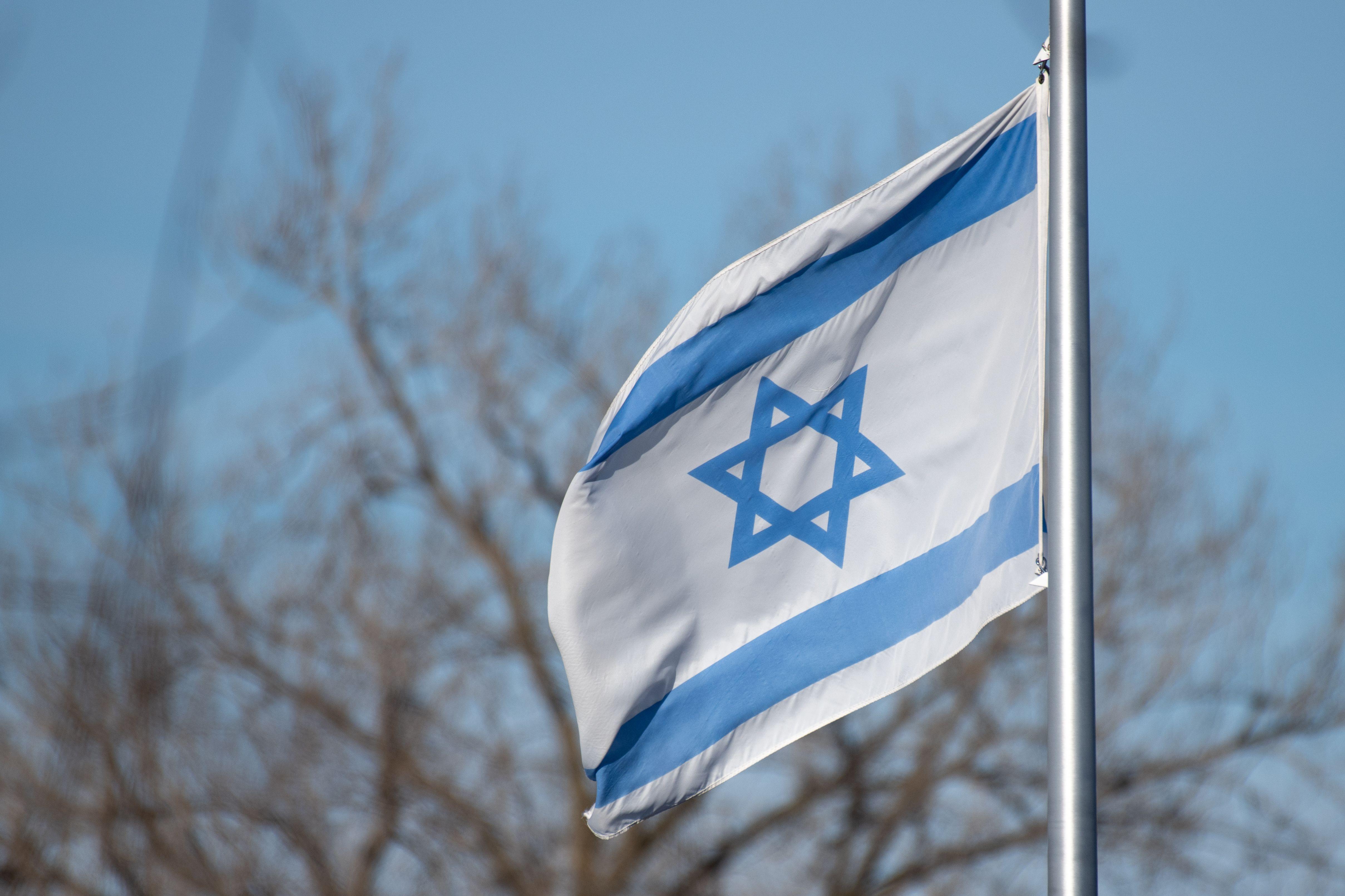 The Israeli flag featuring the Star of David against a clear blue sky and trees.