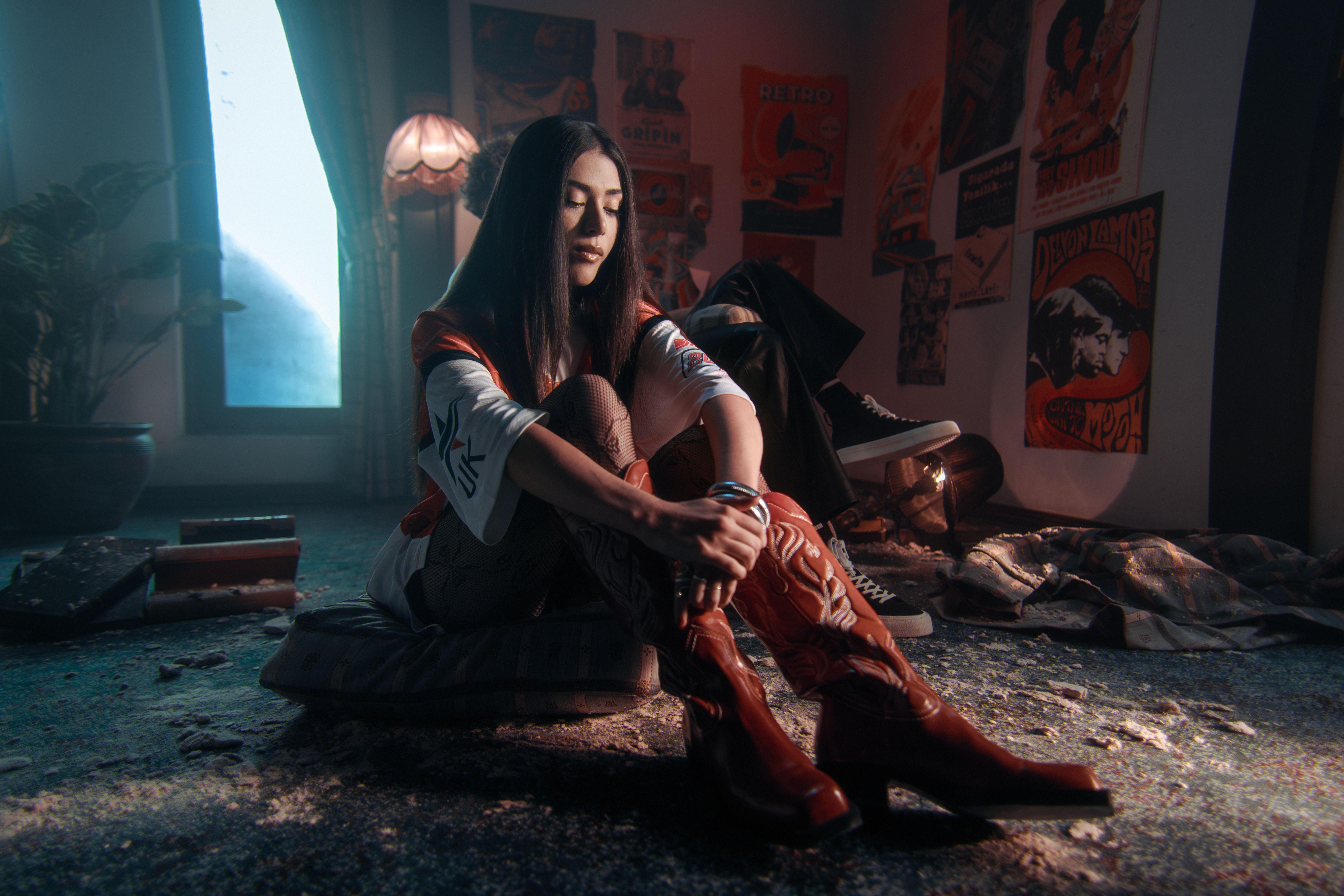 Woman sitting on floor in retro-style room with posters and dim lighting, reflecting nostalgia.