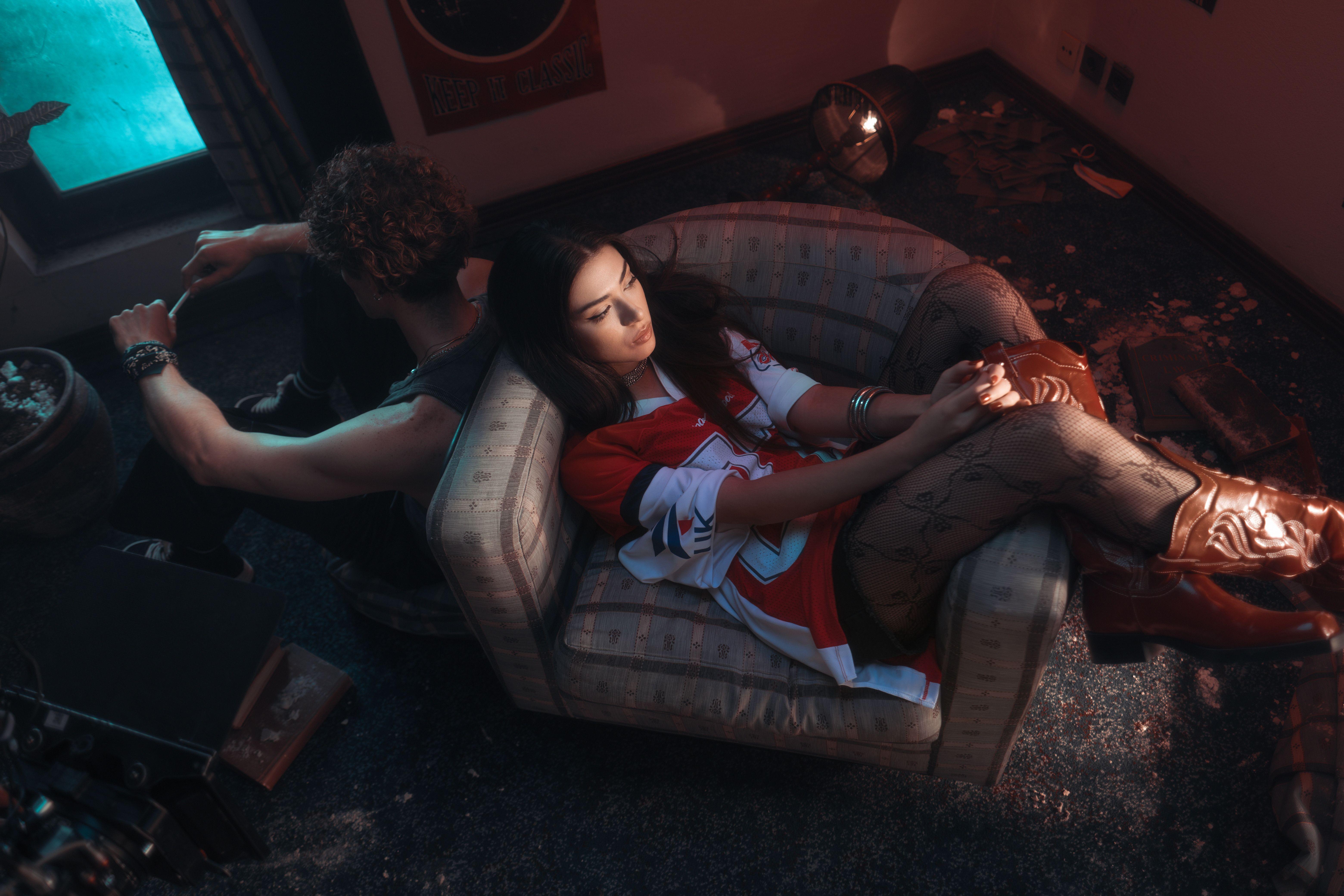 Two young adults in a moody room, both sitting back to back, one on a couch.