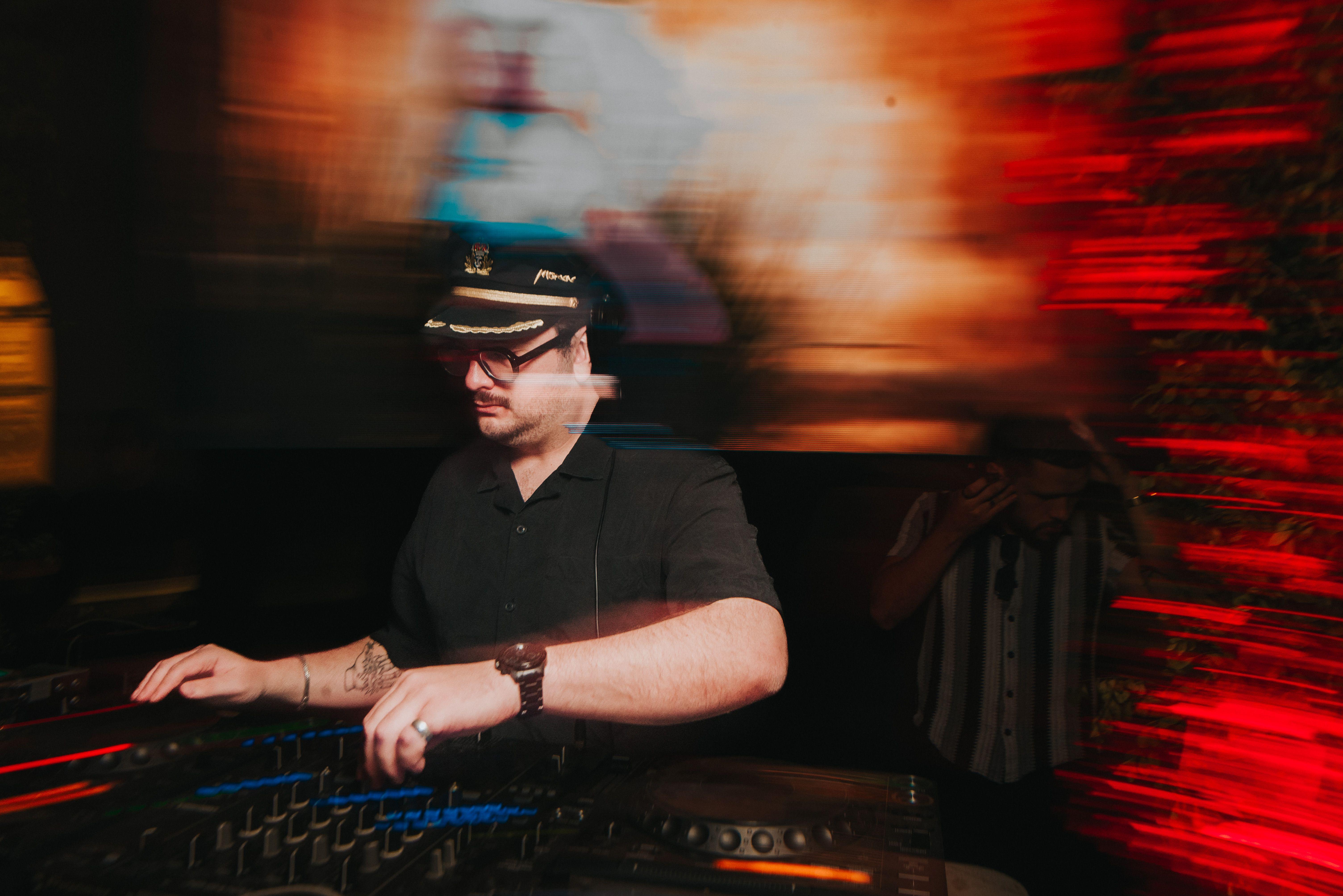A DJ in a captain's hat performs amid vibrant, blurred lights at a nightclub.