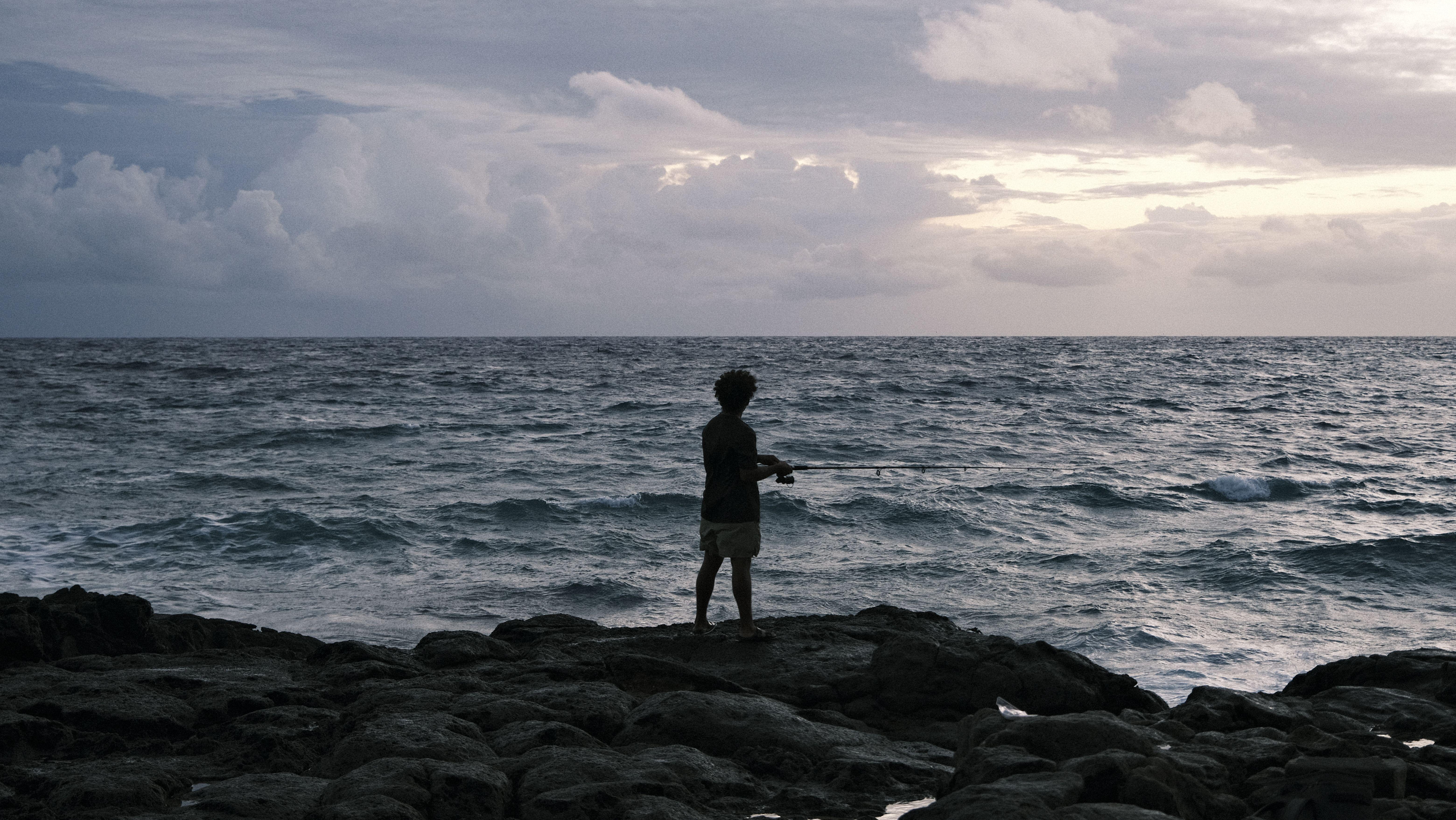 A man fishes alone on a rocky ocean shore under cloudy skies, at dusk.