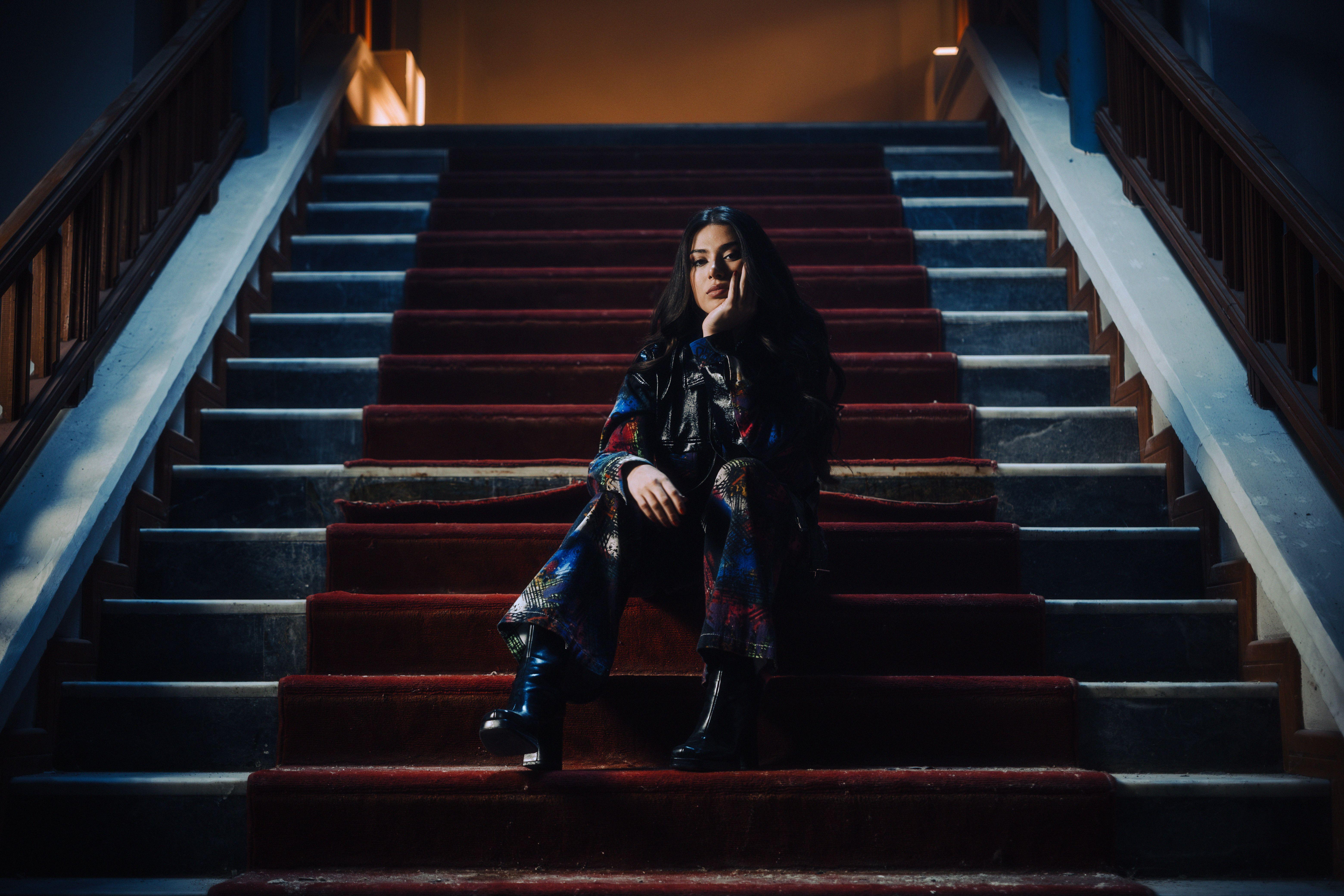 Stylish woman sitting on a red carpeted grand staircase in a moody, theatrical atmosphere.