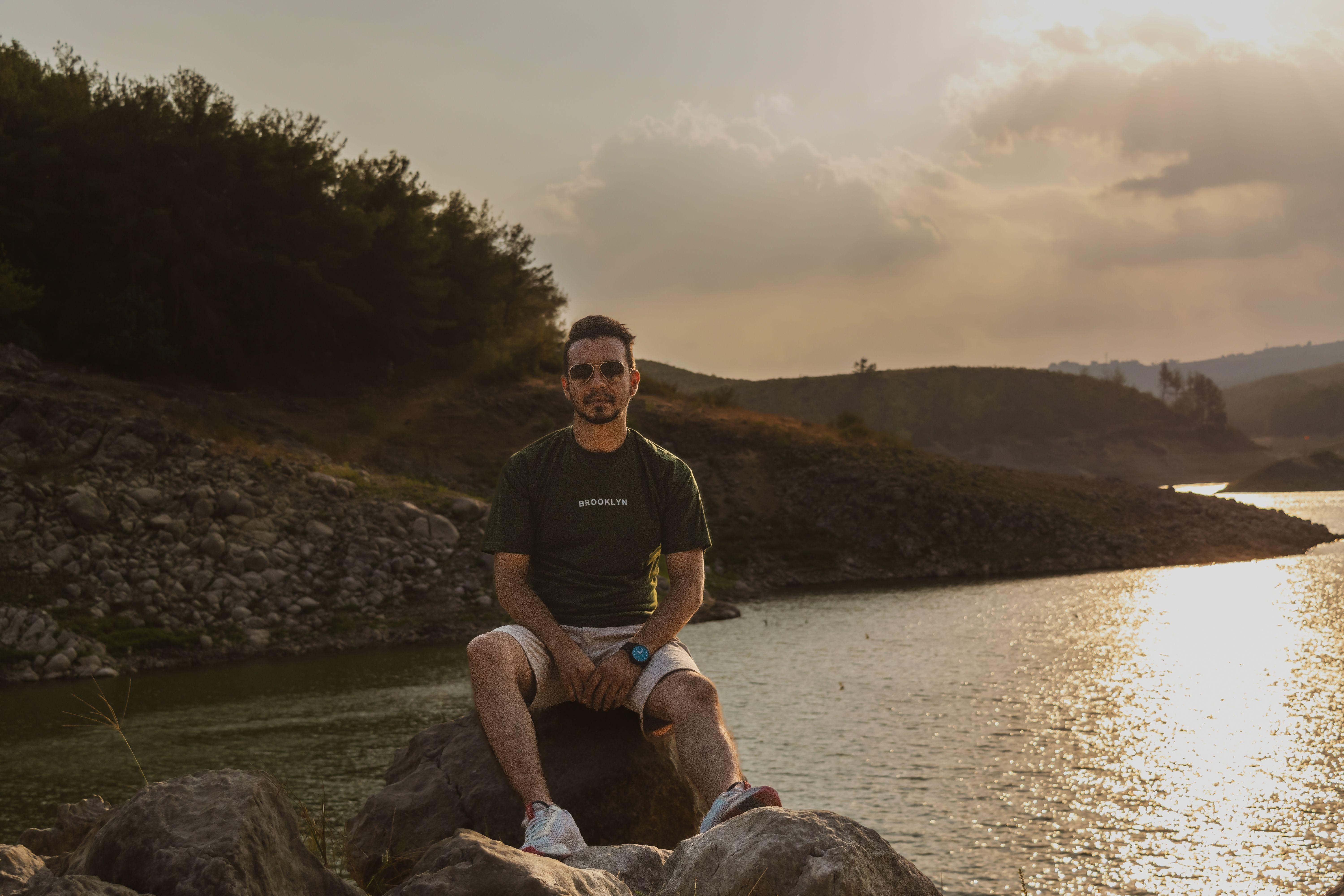A man sits on rocks by a tranquil lake during sunset, exuding calmness and reflection. Ideal for relaxation and travel themes.