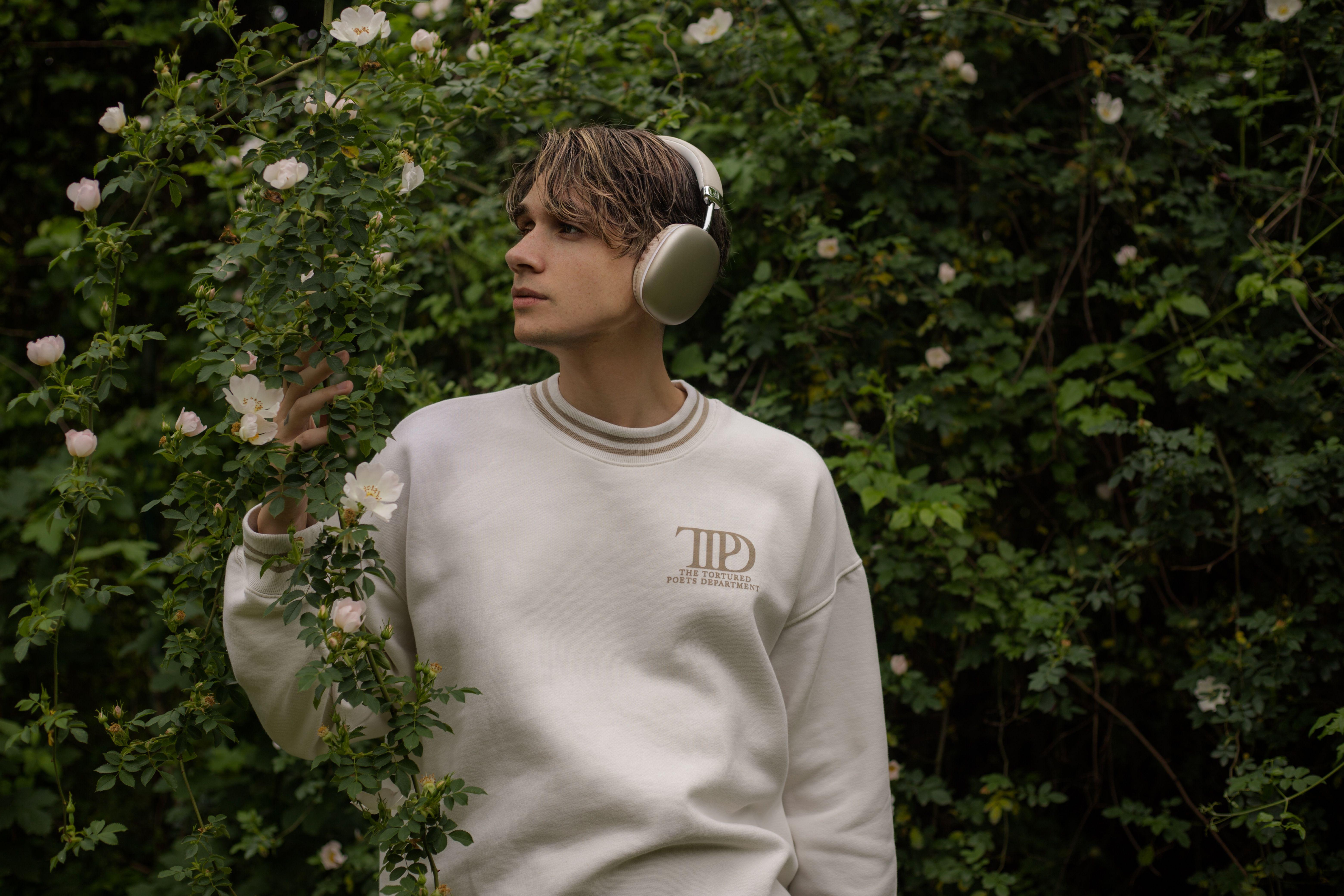 A young man wearing headphones and a white sweatshirt stands amidst lush greenery and flowers.