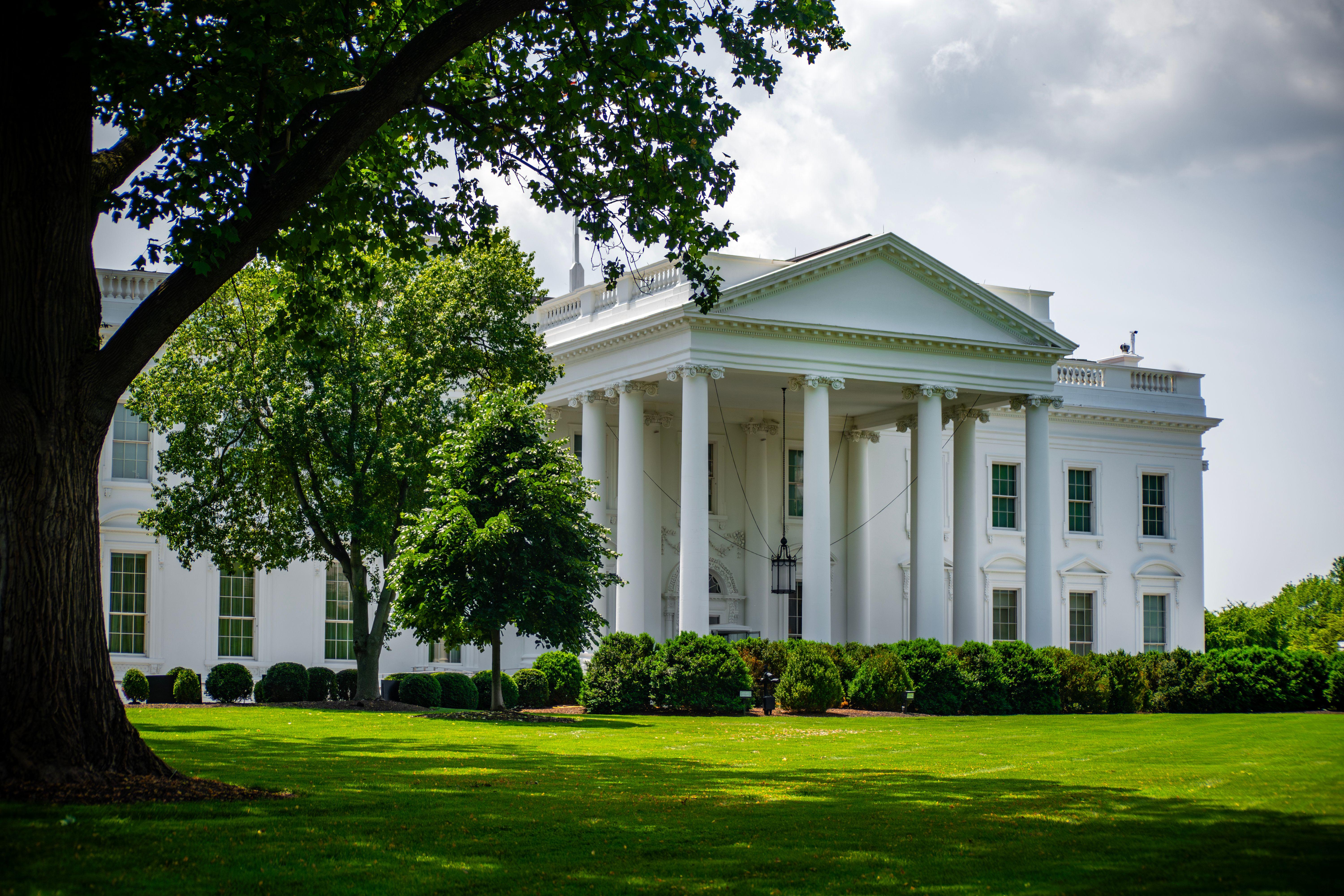 The White House framed by trees and greenery, in Washington, D.C., under a bright sky.