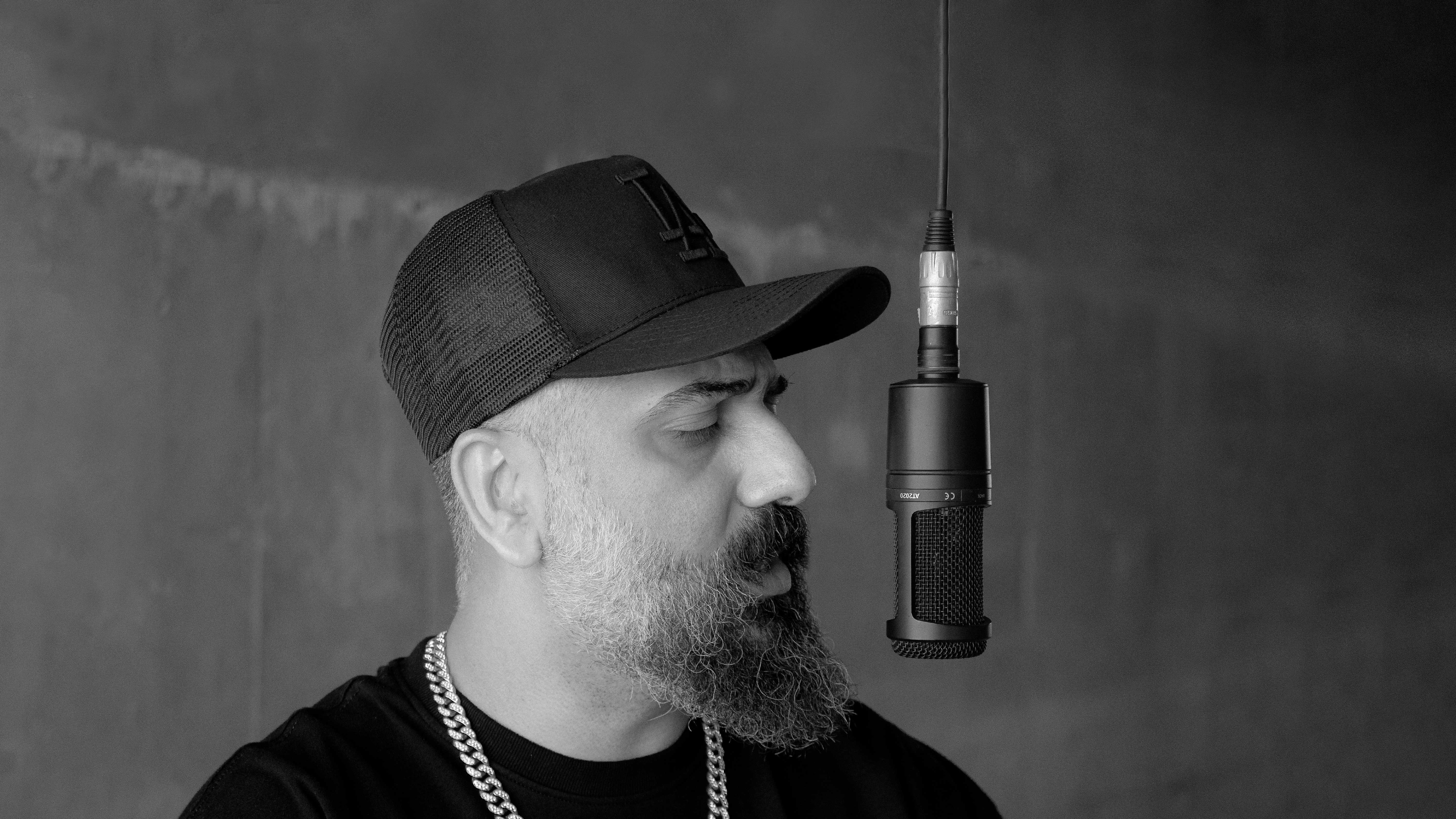 Black and white portrait of a bearded rapper wearing a cap in front of a microphone.