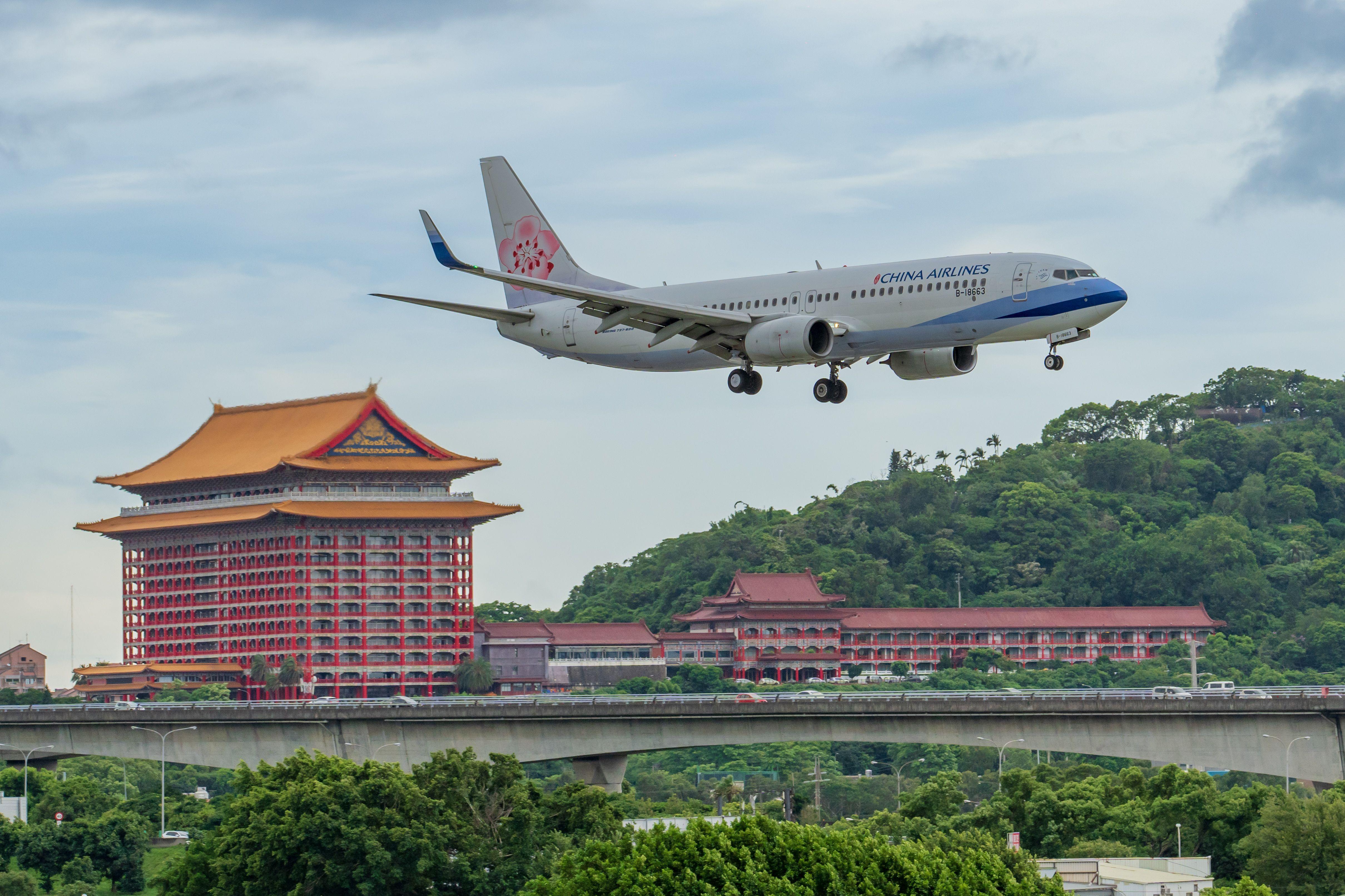 China Airlines plane approaches landing over Grand Hotel Taipei amidst lush greenery and cloudy sky.