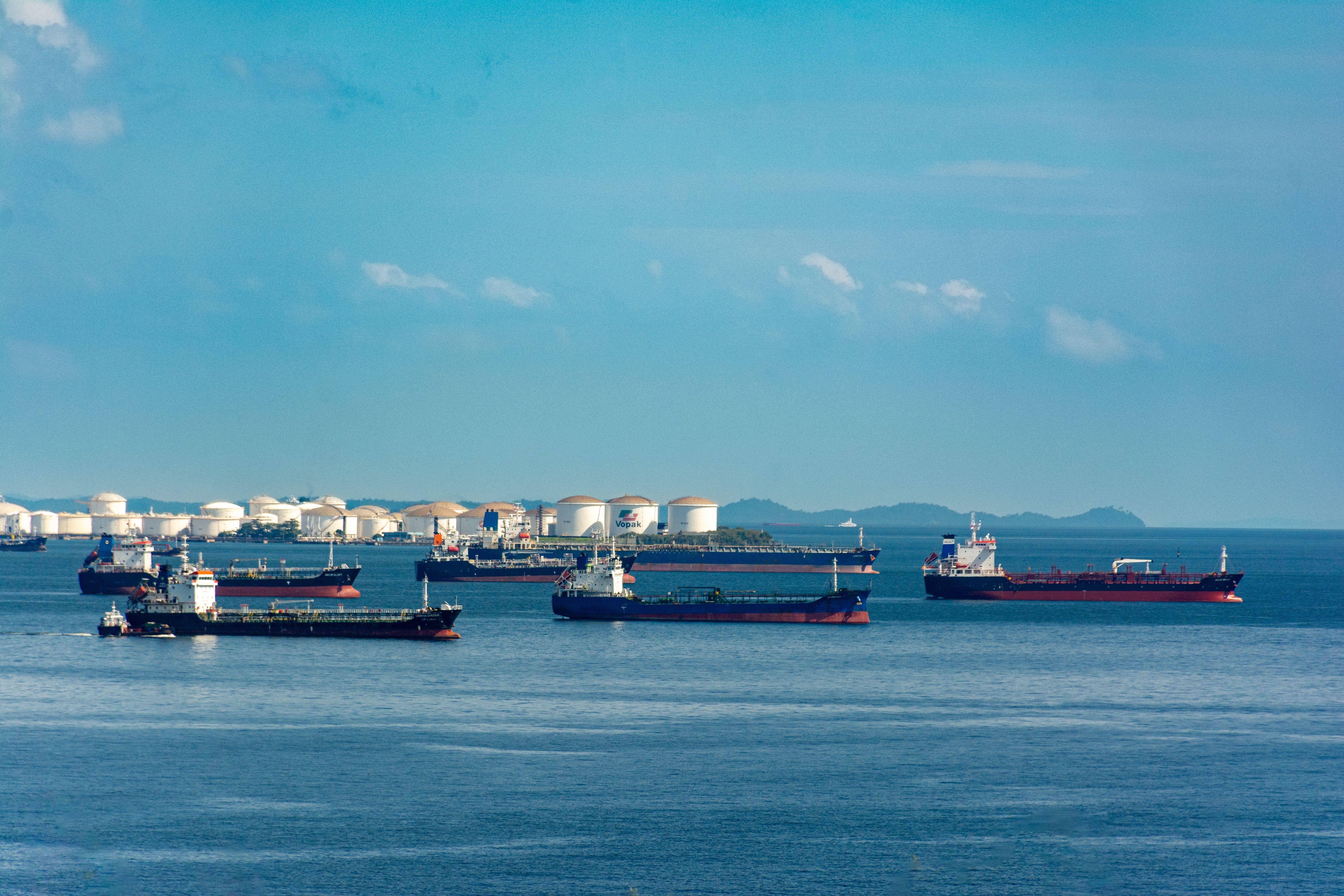 A fleet of cargo ships docked near oil storage tanks along a serene coastline with a clear blue sky above.
