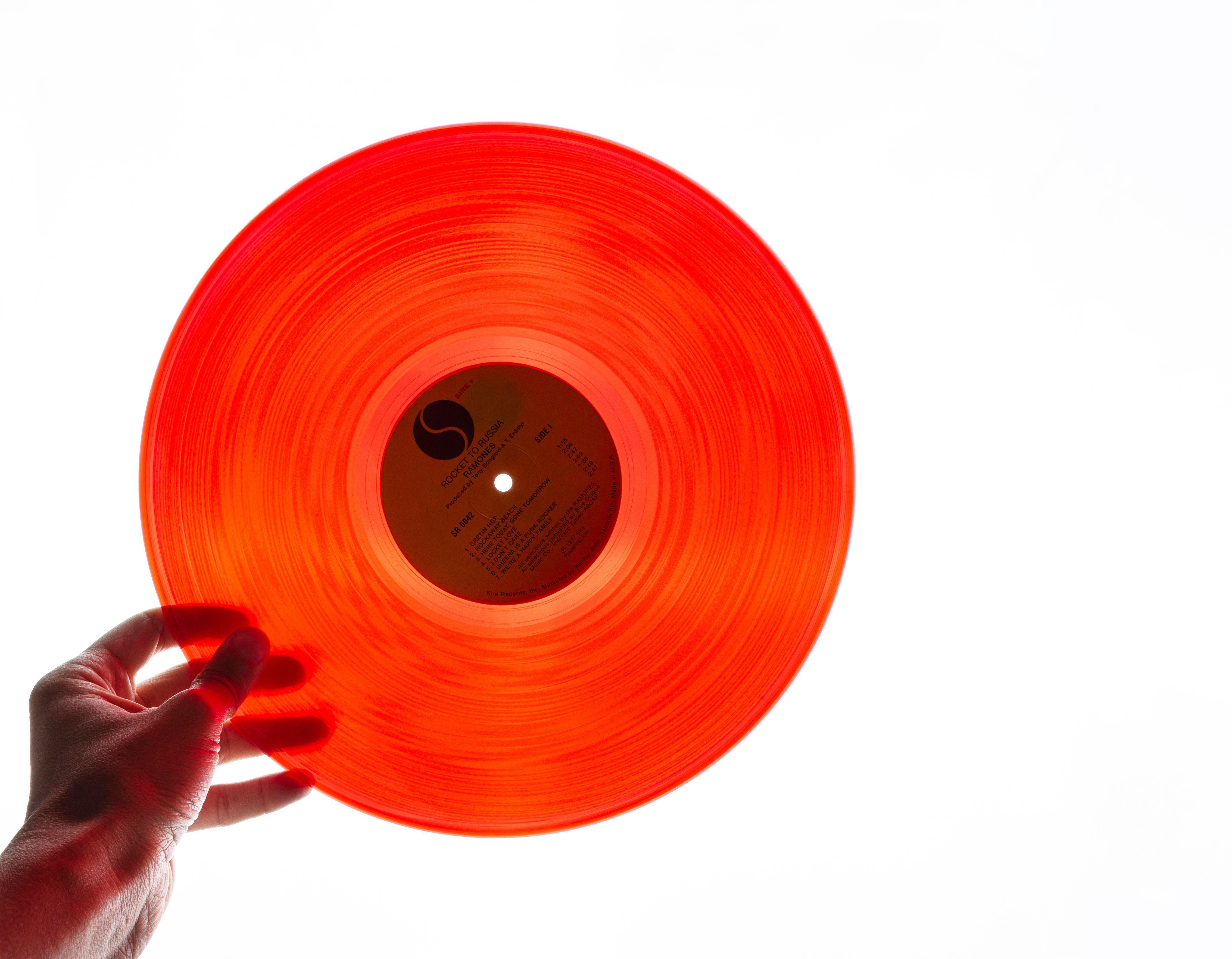 A bright orange vinyl record being held by a hand against a plain white background, showcasing its translucency.