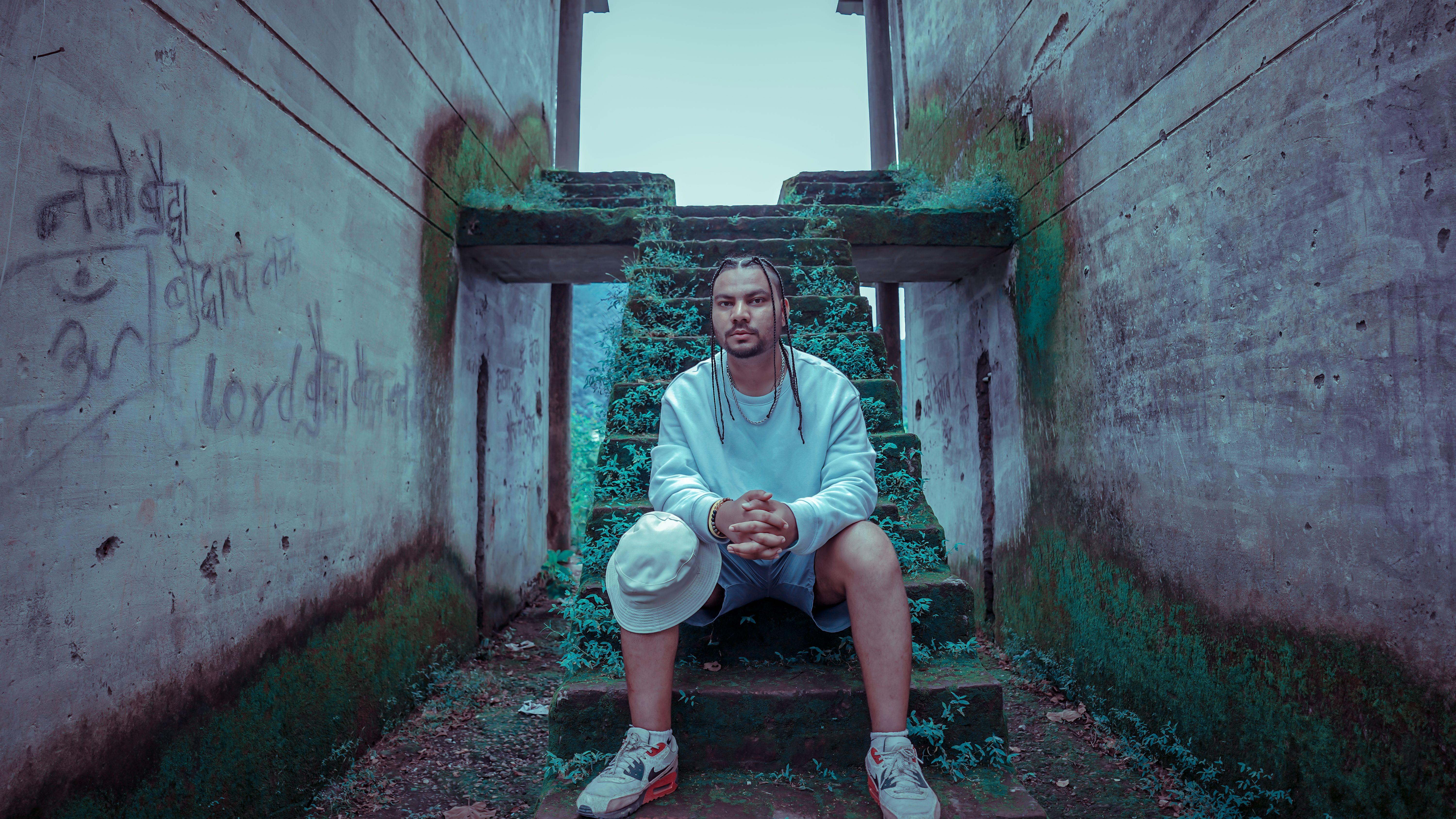 A stylish young man sits on mossy steps in Rishikesh, India, showcasing urban fashion.