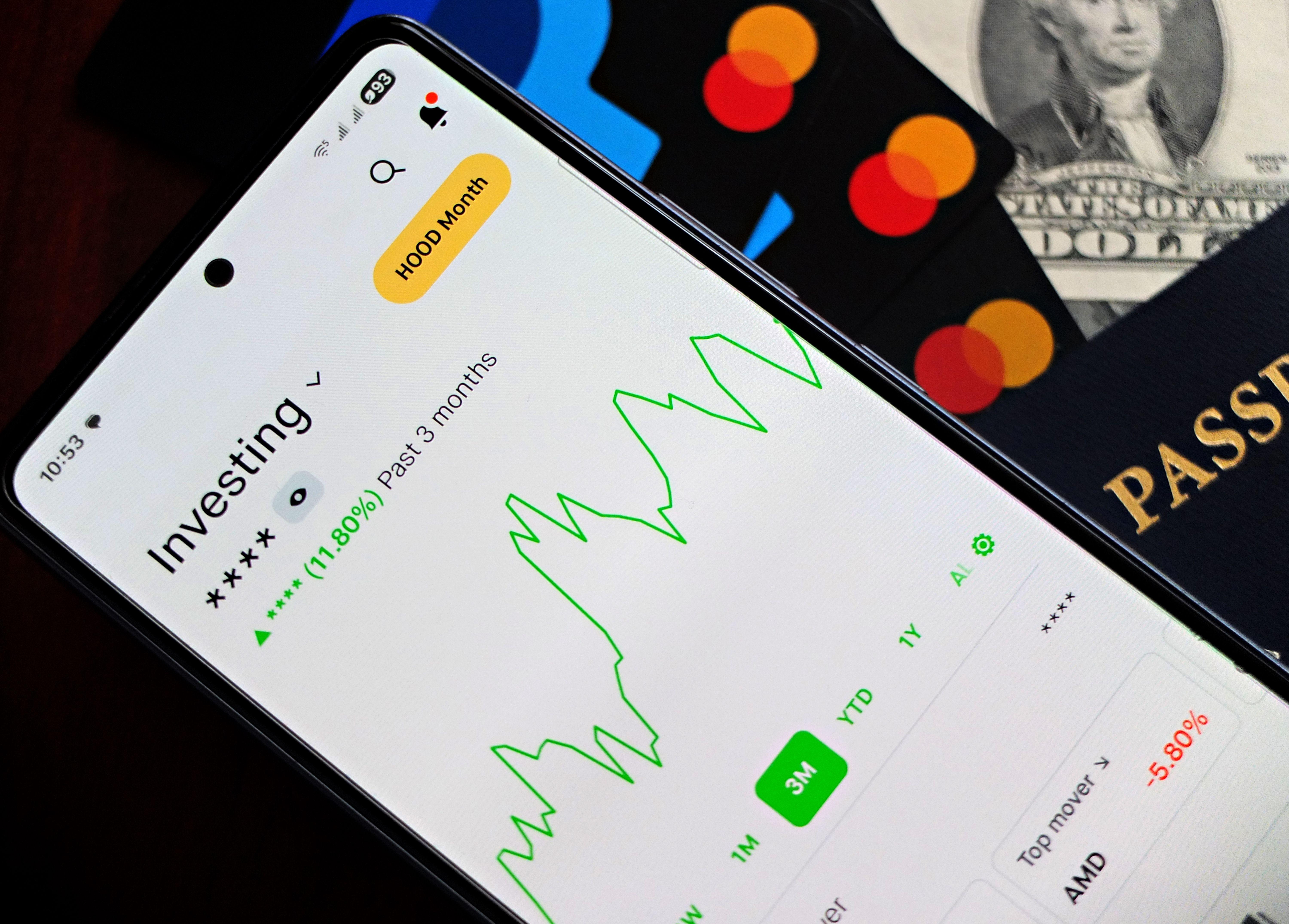 A smartphone showing an investment app with green growth indicators, surrounded by credit cards, US dollars, and a passport.
