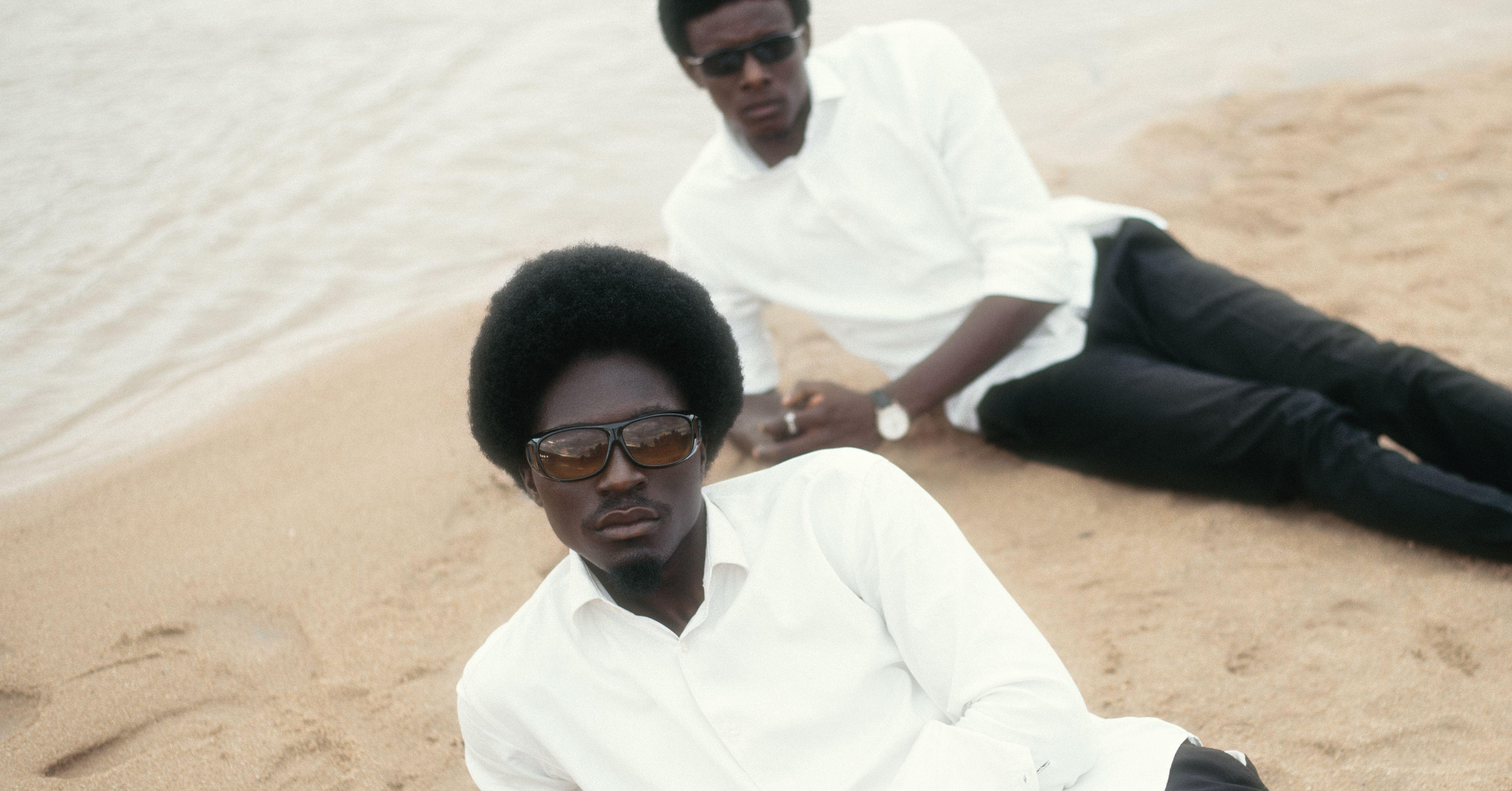 Two stylish men in sunglasses relax on a sandy beach in minimalistic attire.