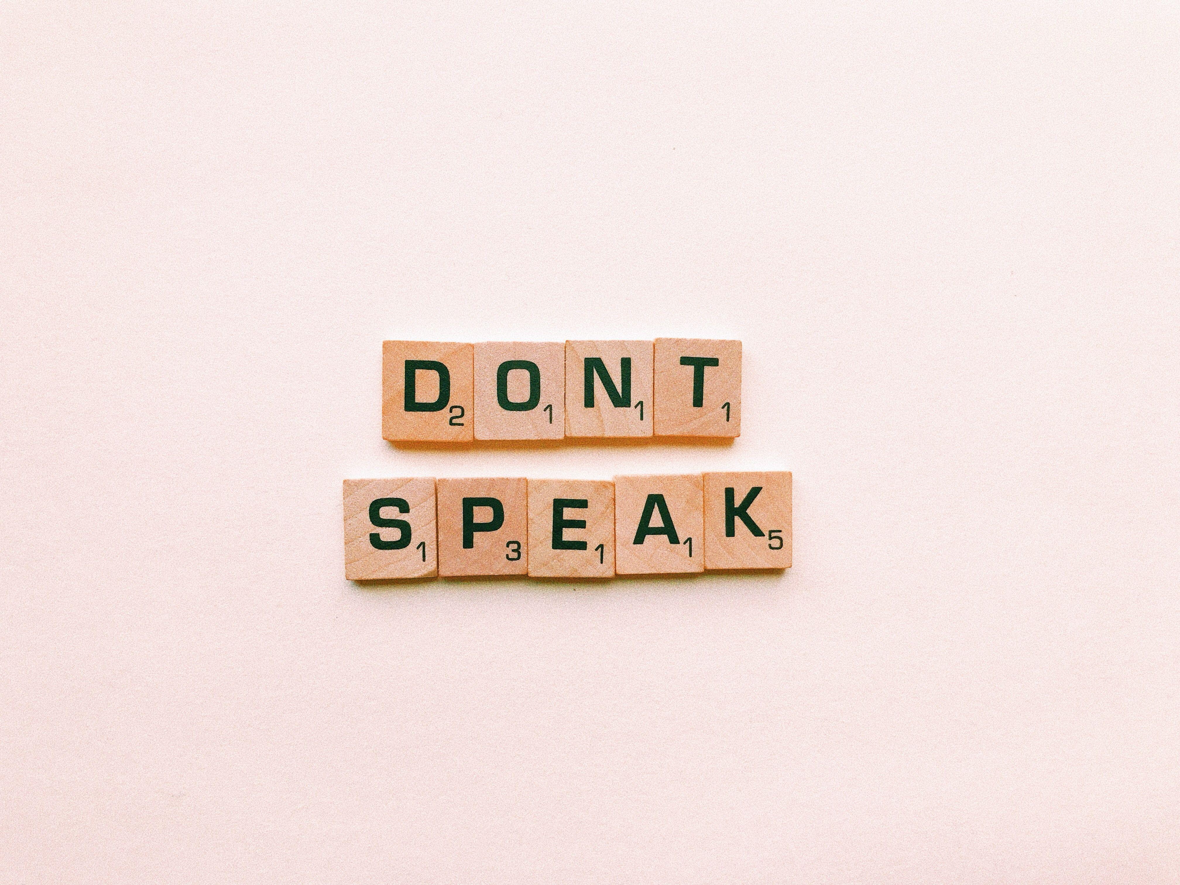 Wooden Scrabble tiles spelling 'Don't Speak' on a pink background, emphasizing silence.