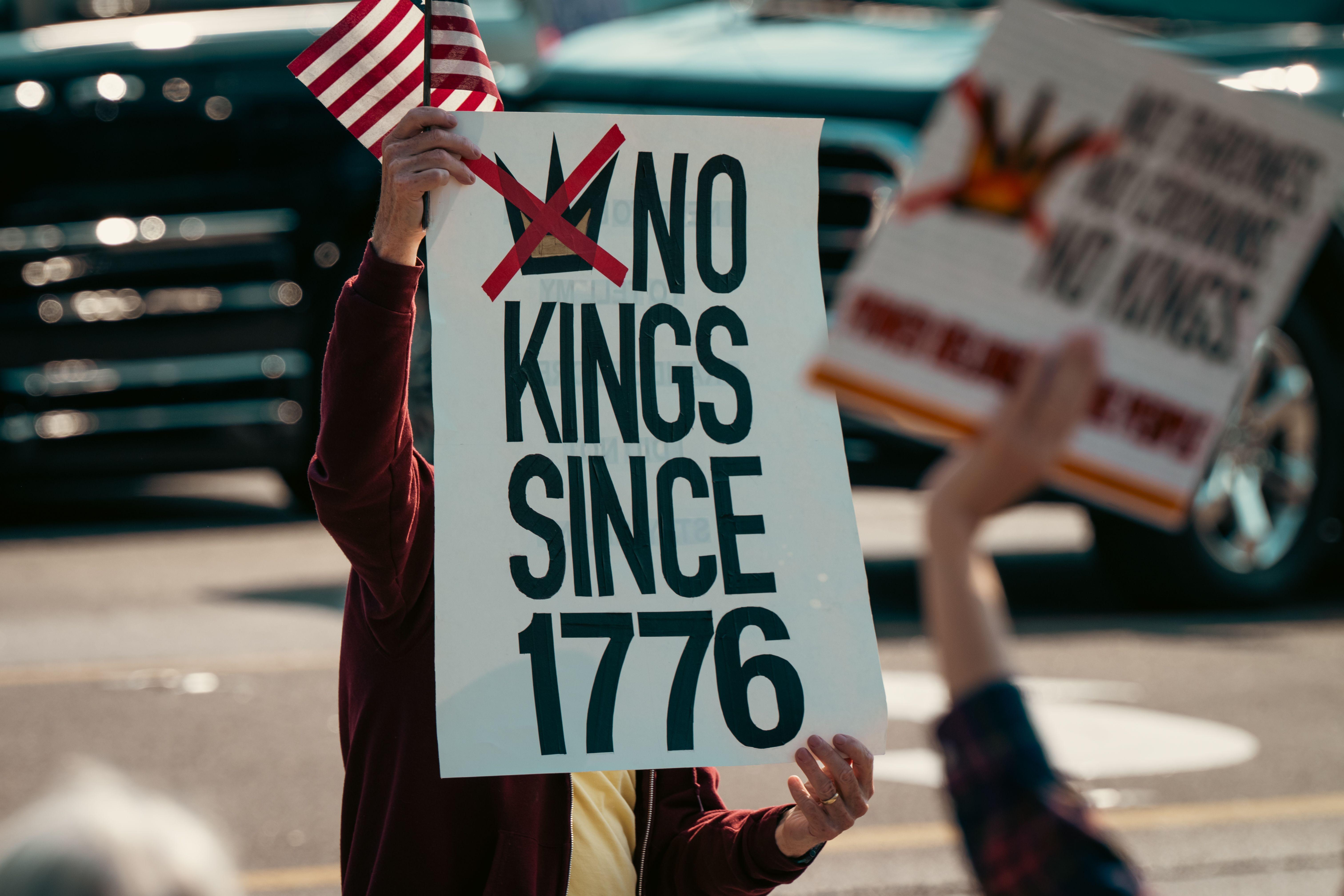 Protest sign displaying 'No Kings Since 1776' held alongside a small US flag, emphasizing democratic sentiments.