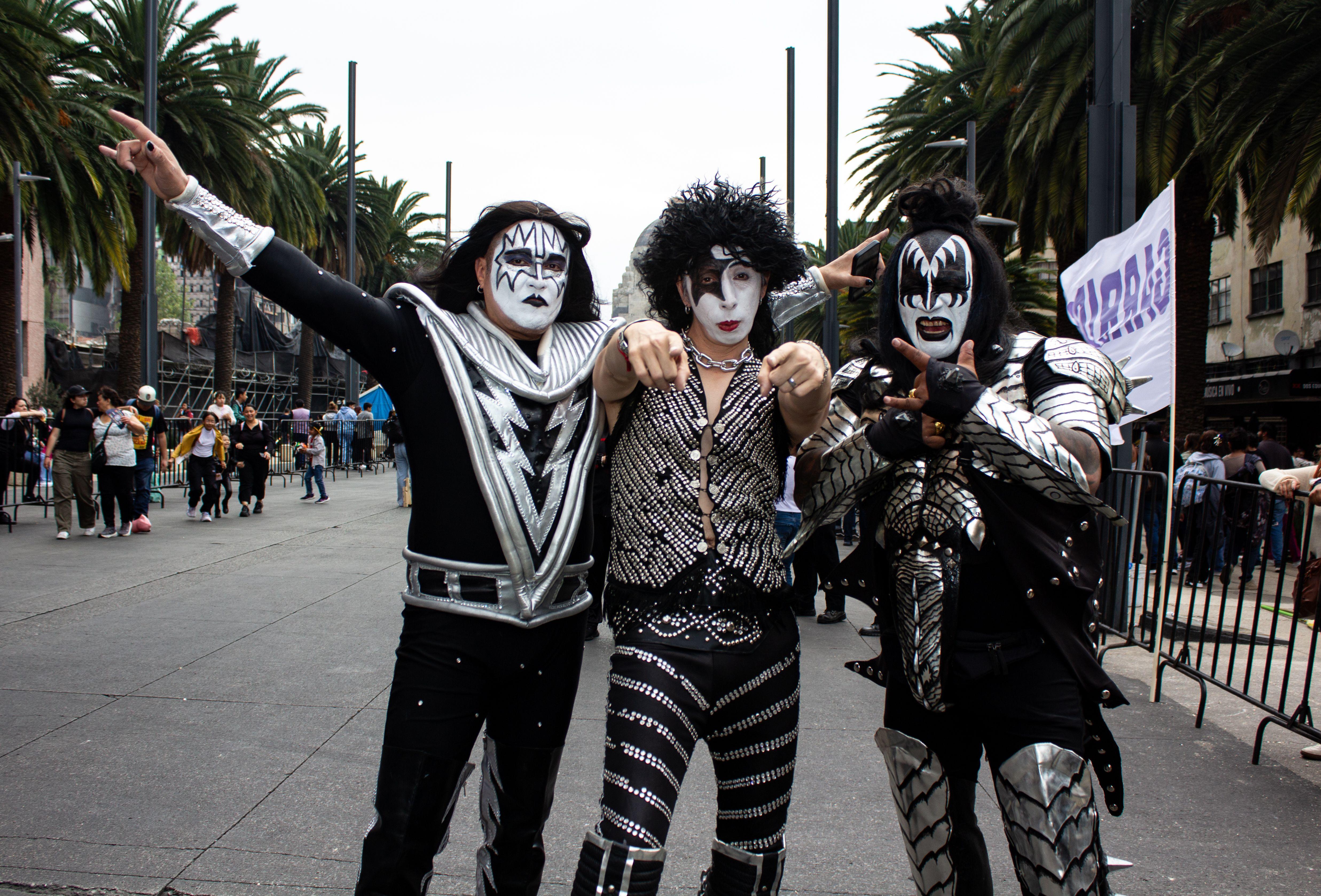 Three adults dressed as KISS band members pose in a lively street scene in Mexico City.