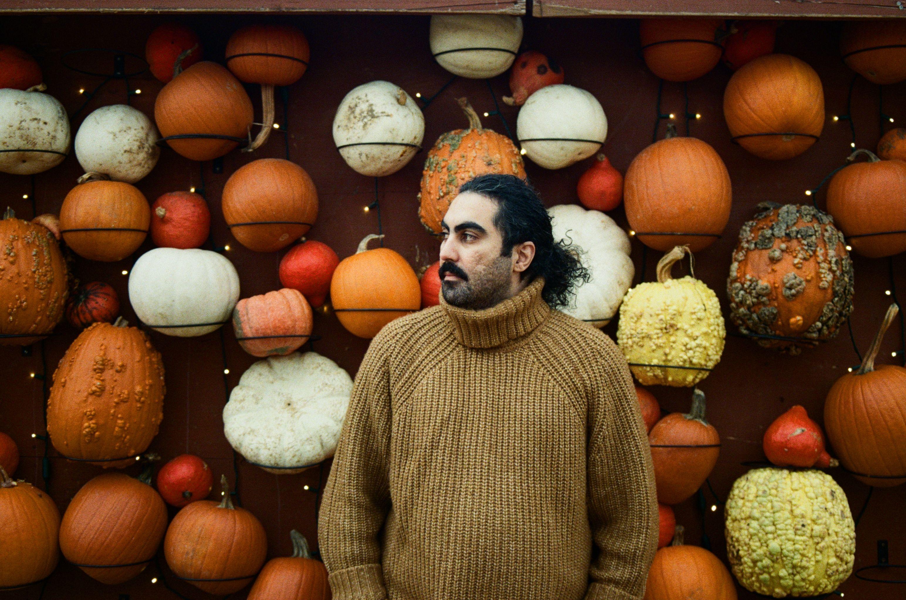 Man in a warm sweater stands against a colorful pumpkin wall, showcasing autumn vibes.