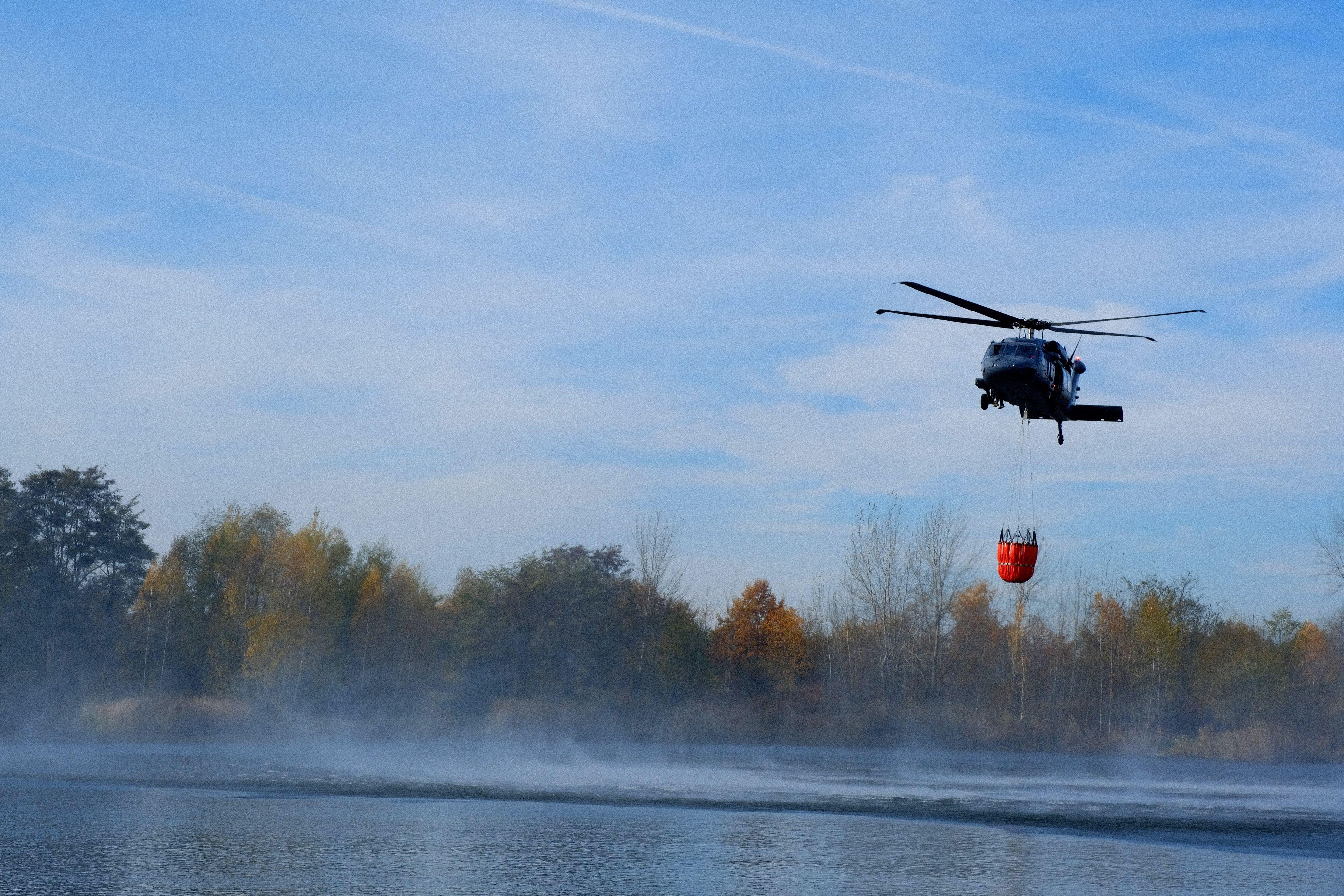 A helicopter gathers water from a lake for firefighting efforts. Blue sky and autumn trees.