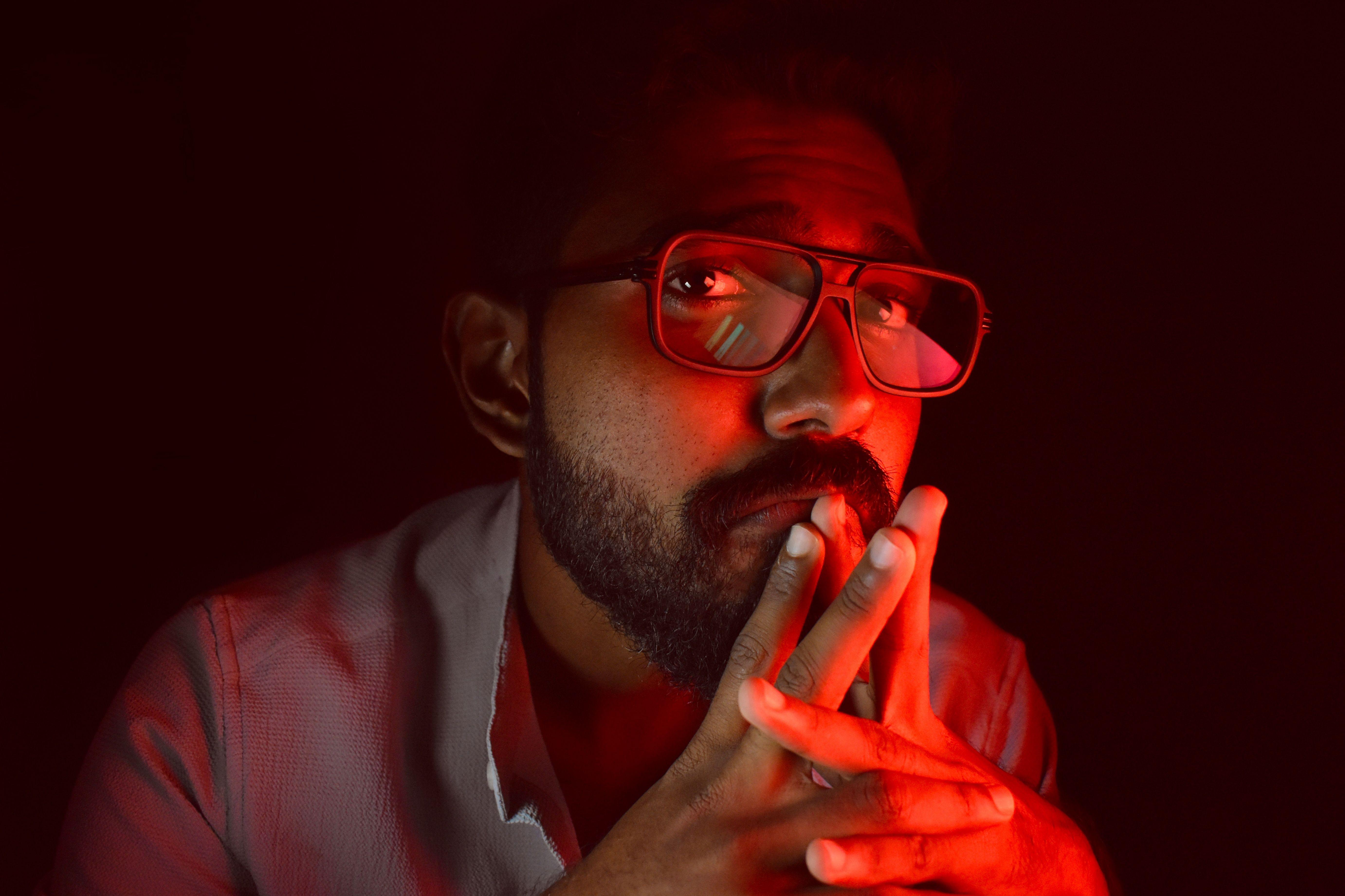 A thoughtful man with red ambient lighting, wearing glasses and a beard, looking upwards.