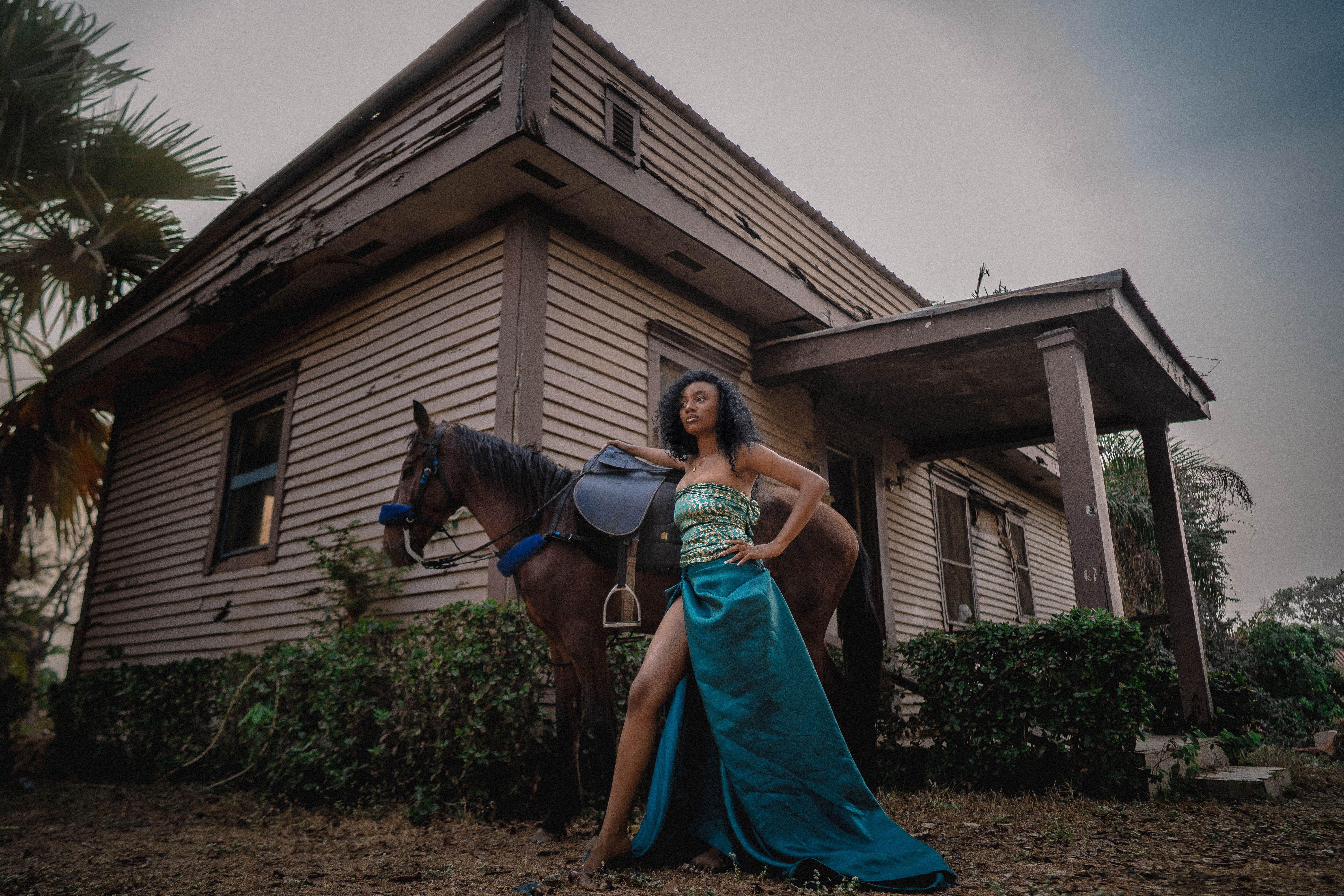 A woman in a glamorous dress poses confidently with a horse beside a rustic wooden house.
