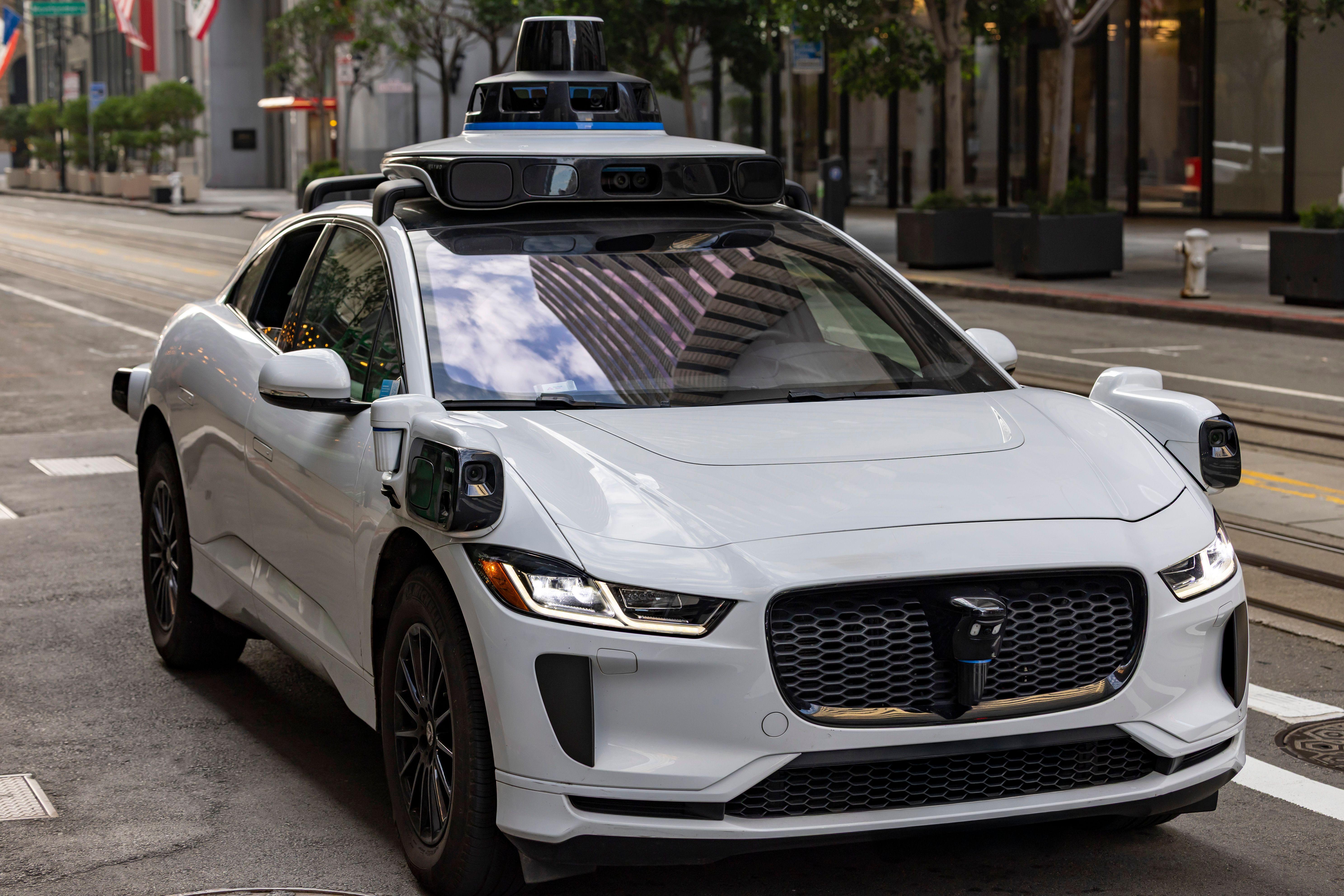 A white autonomous vehicle navigating a city street, reflecting urban architecture in daylight.