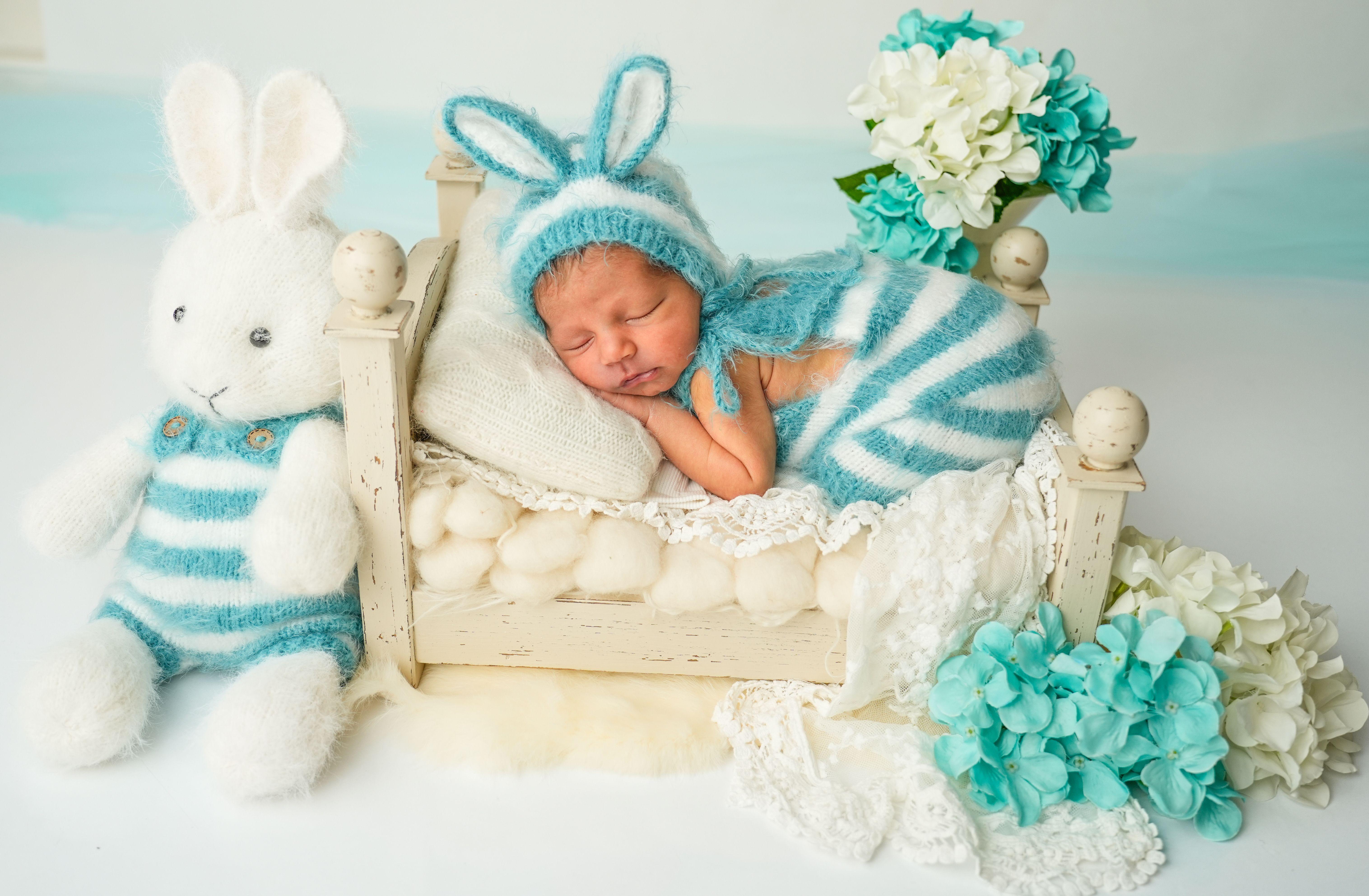 Newborn baby wearing a blue bunny costume asleep on a bed with flowers and a stuffed toy.