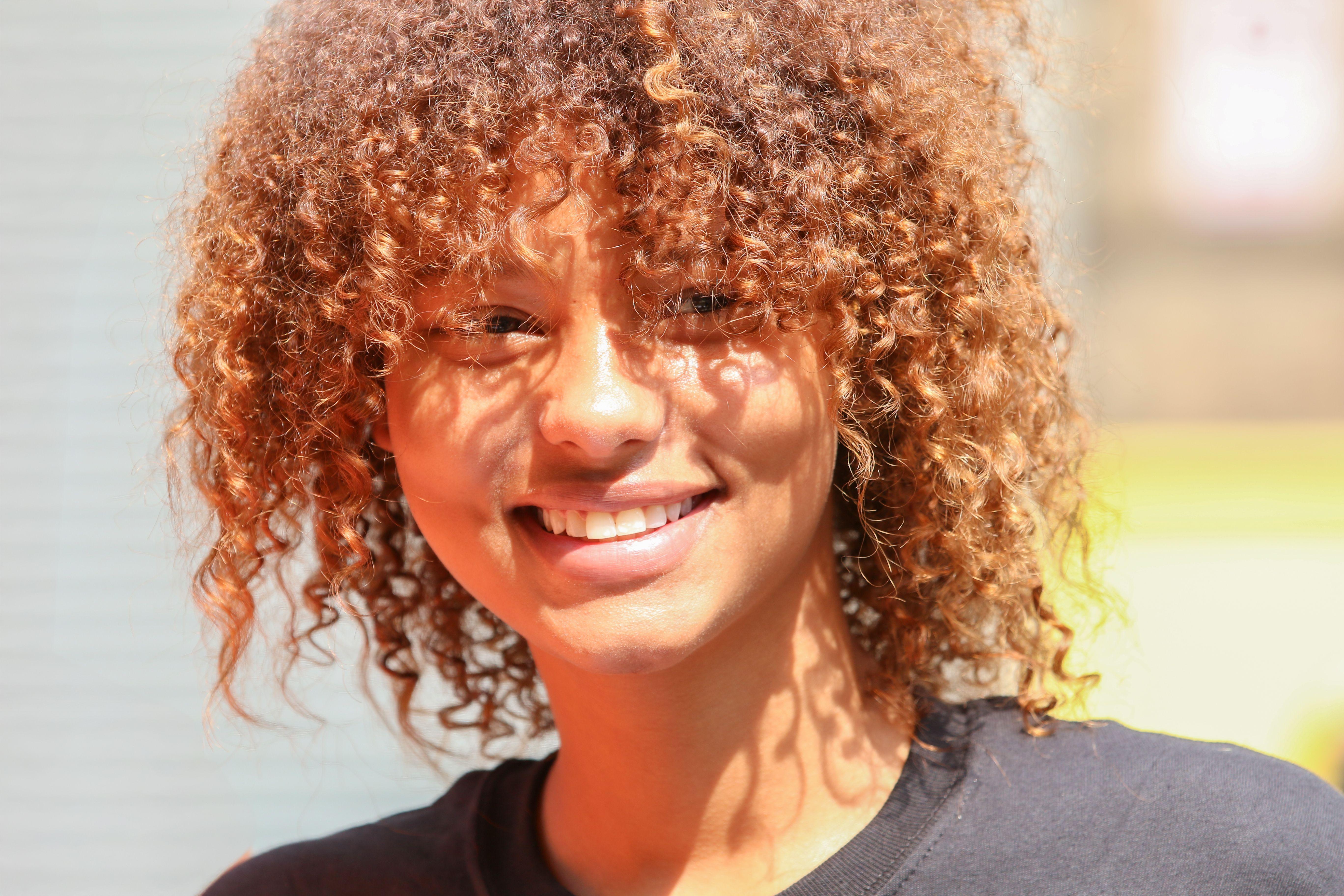 A young woman with curly hair smiles brightly during a sunny day outdoors, radiating joy and warmth.