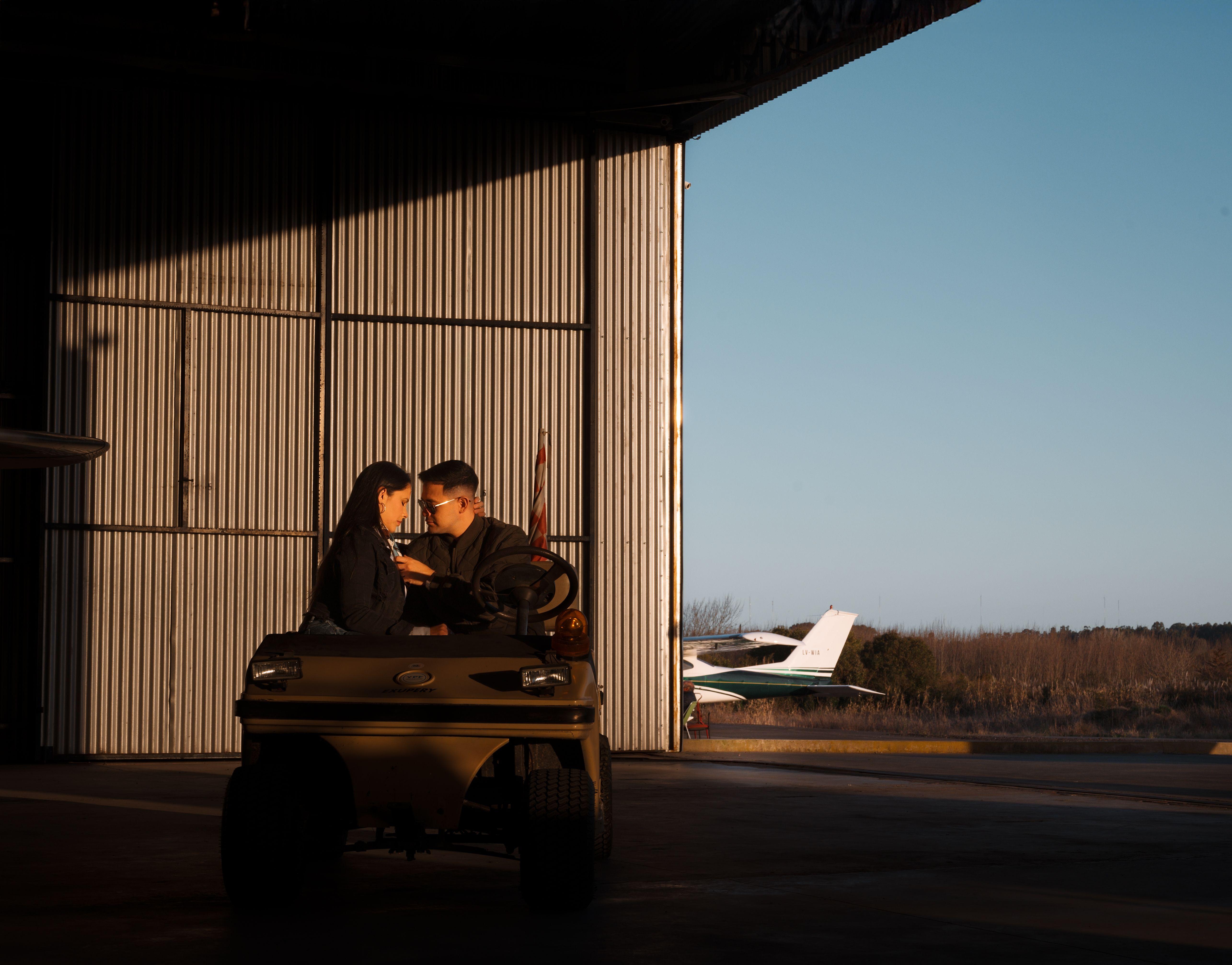 A couple sits inside a hangar, enjoying a moment with an airplane visible outside.