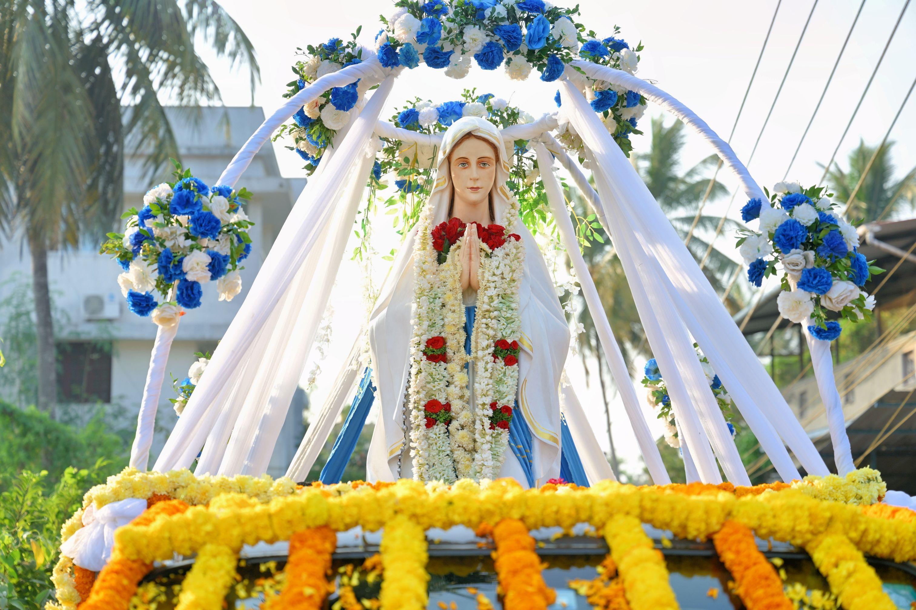 Statue of Our Lady of Lourdes adorned with colorful flowers in an outdoor setting with palm trees.