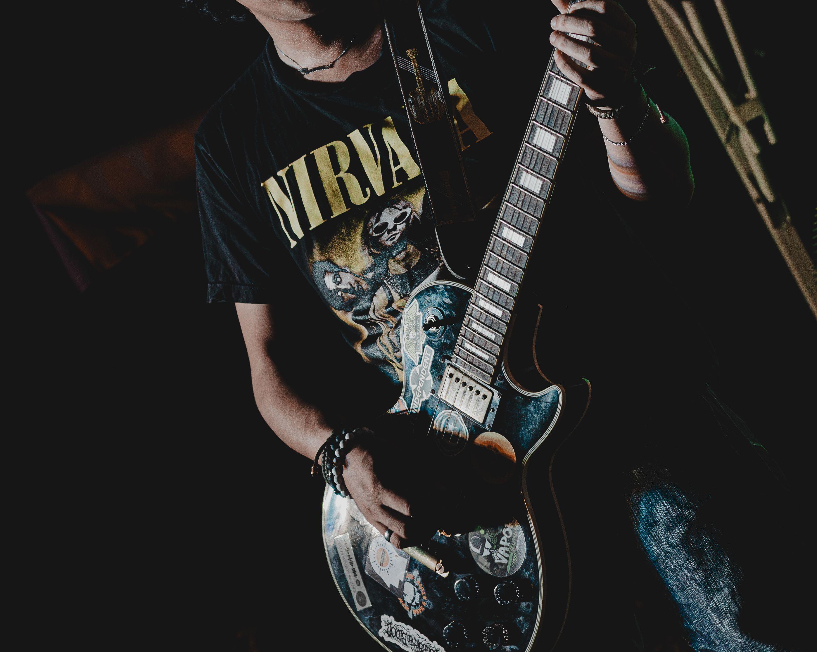 Close-up of a guitarist playing an electric guitar indoors, wearing a Nirvana shirt.
