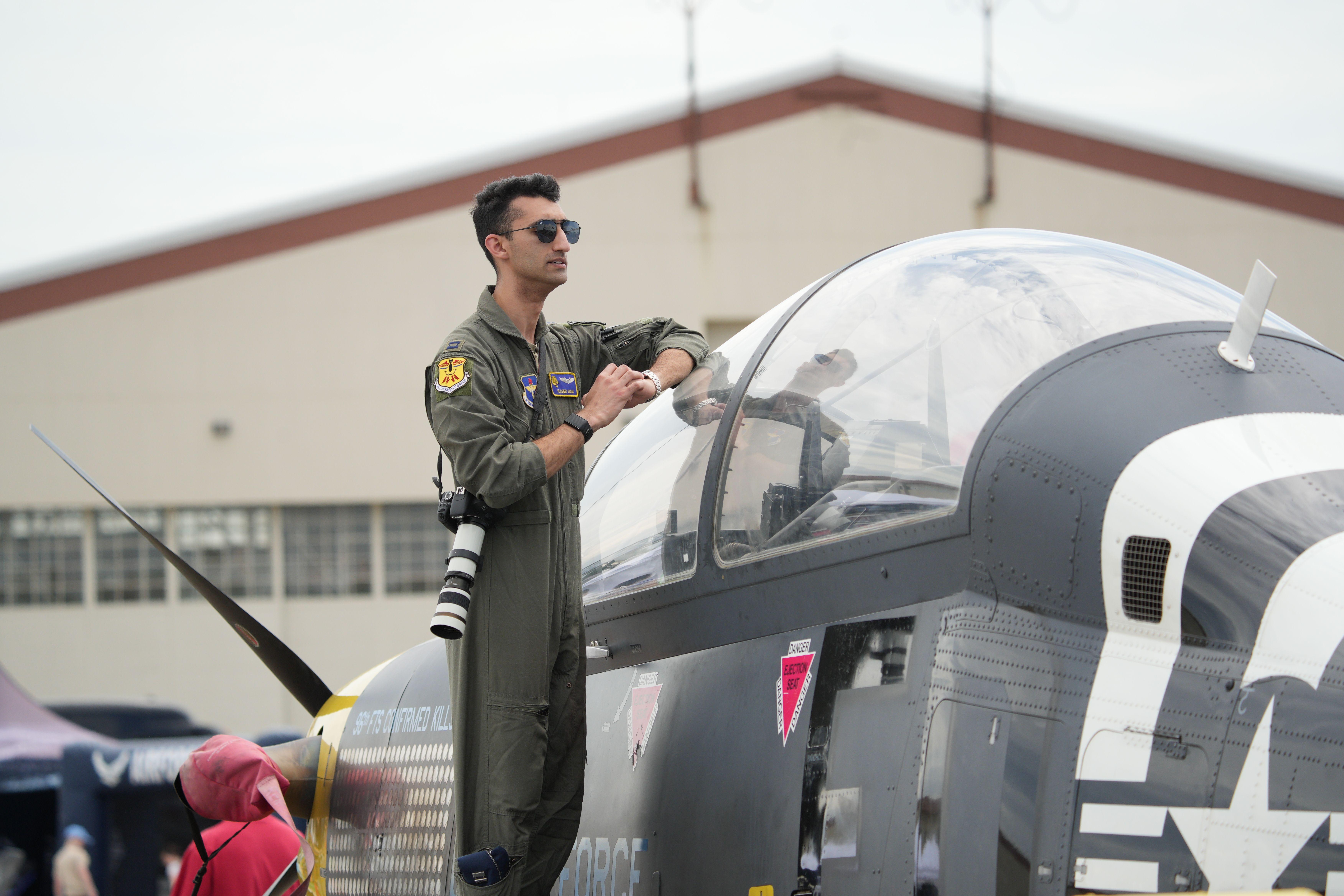 A military pilot in uniform stands confidently atop a modern fighter jet at an airshow.