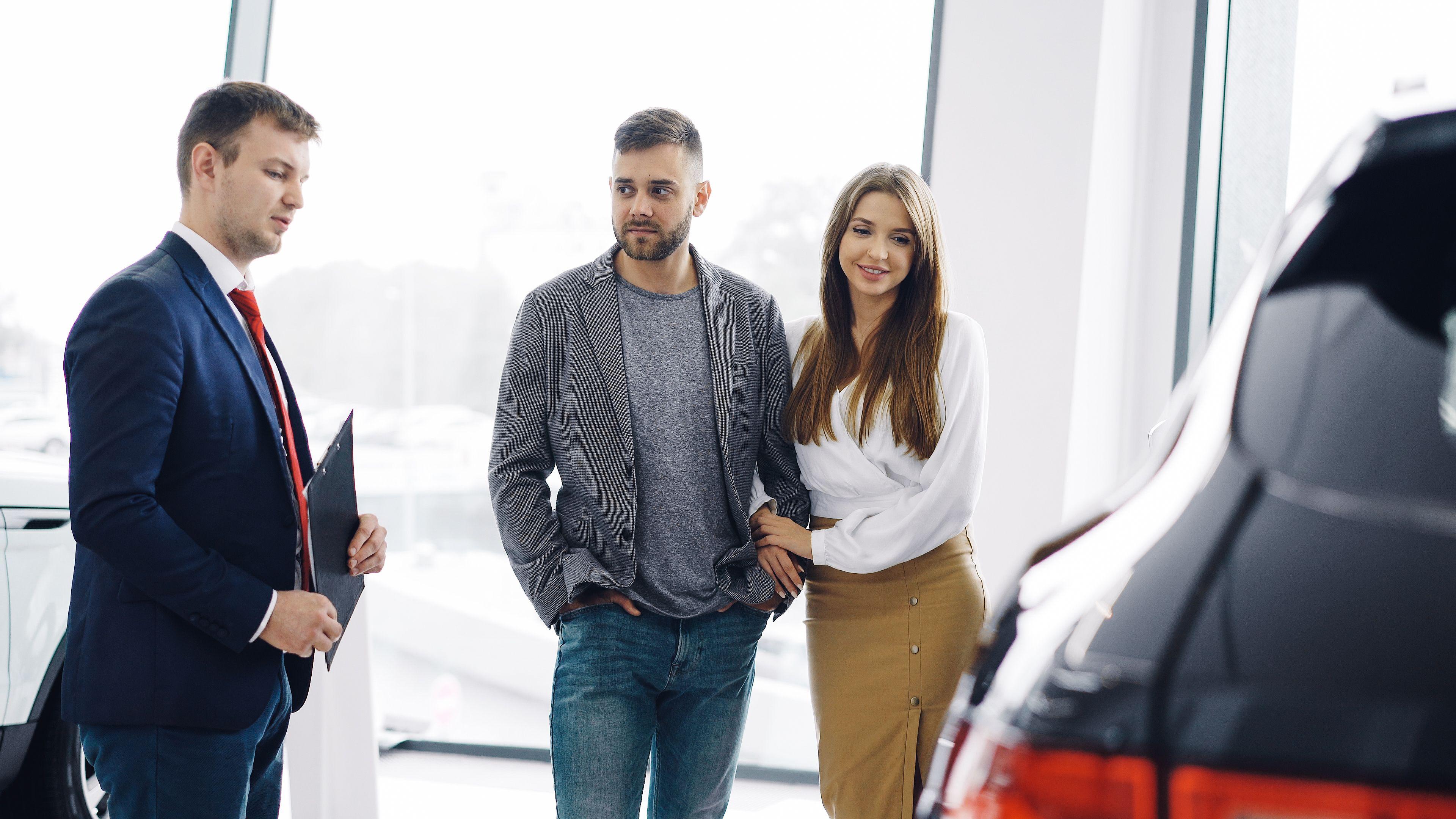 Smiling couple with salesman in car showroom discussing features of a car.