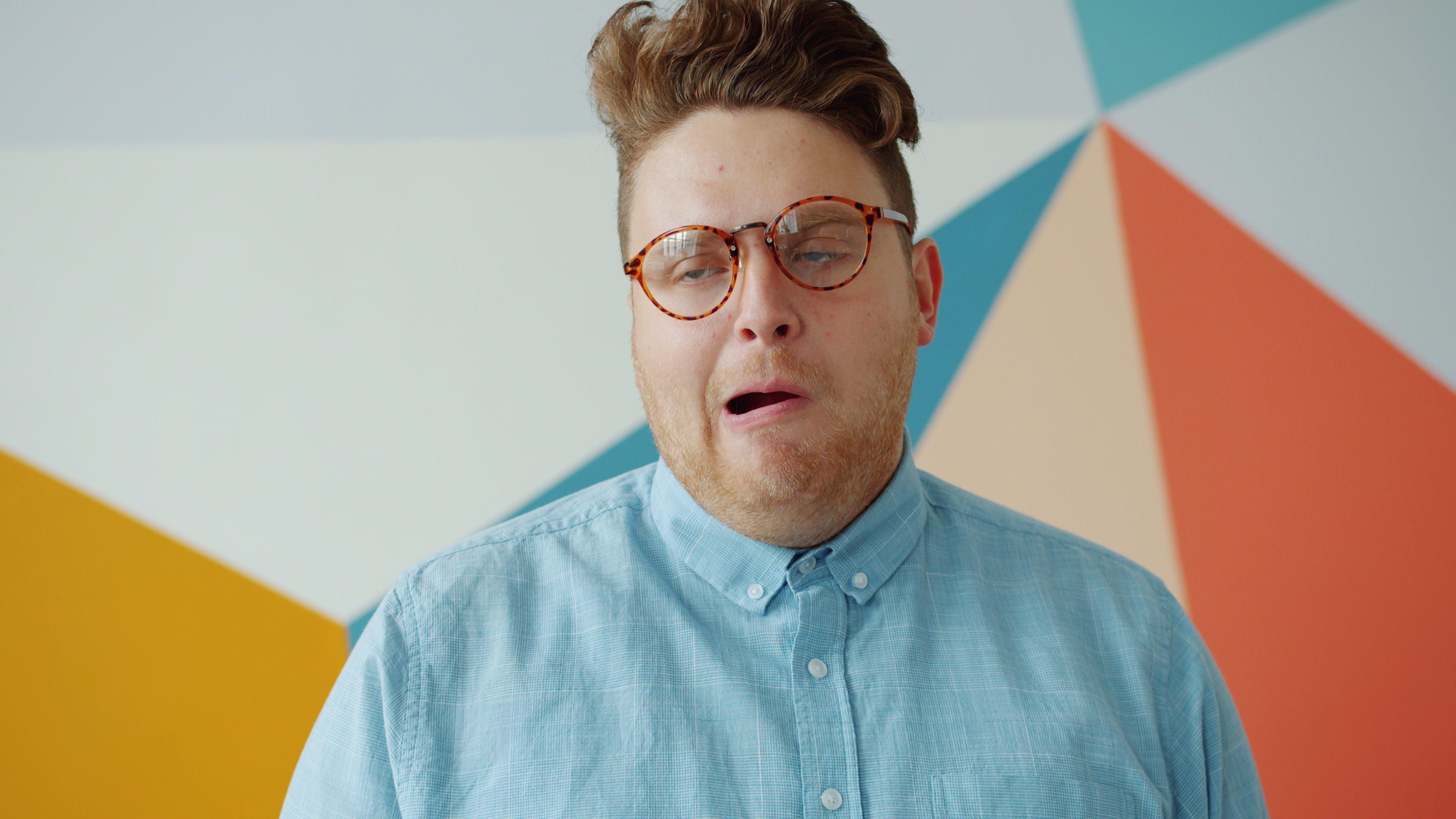 A young man with glasses makes a humorous expression against a colorful geometric wall.
