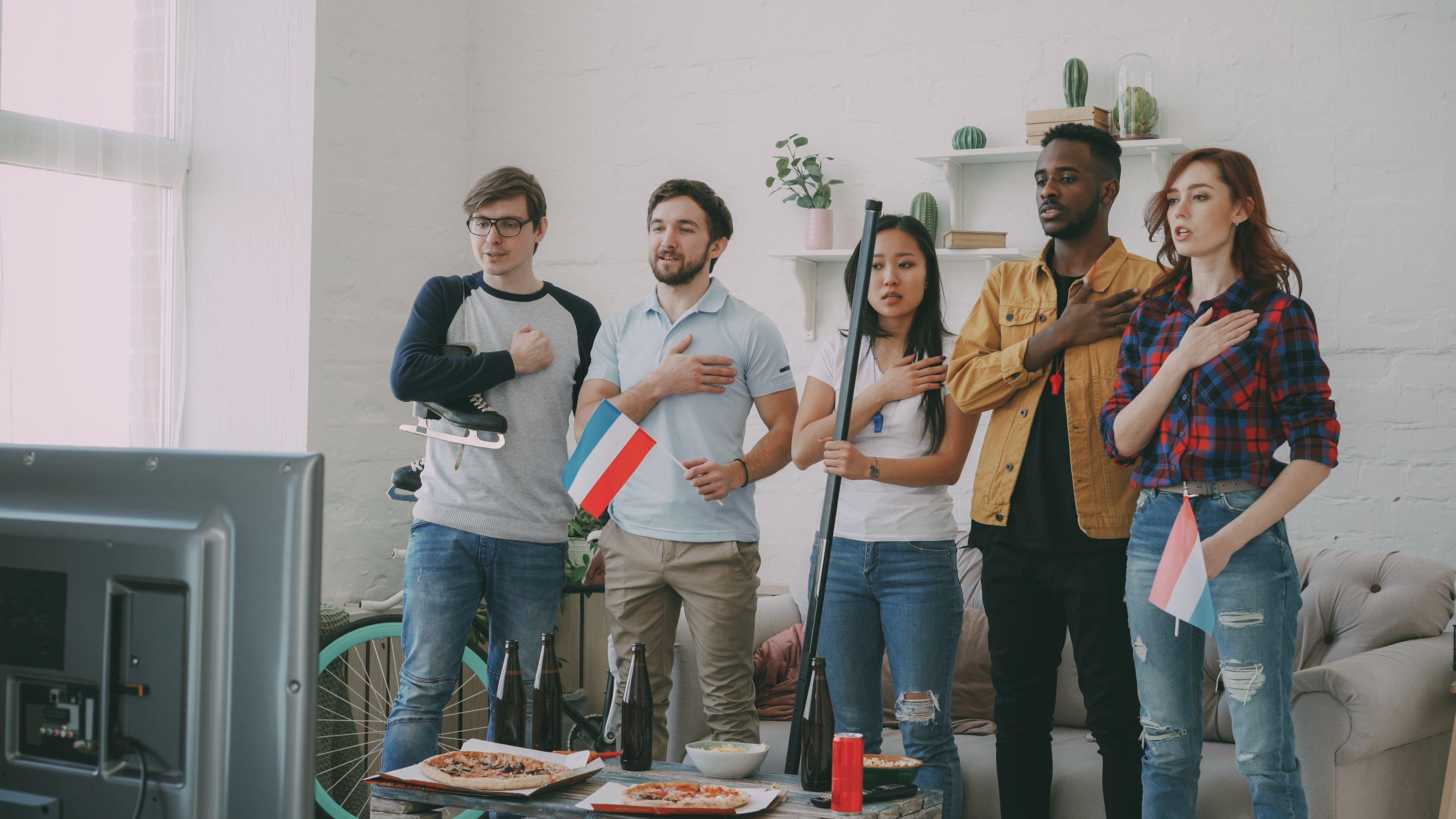 Five friends watching sports, hand on heart, holding flags, showing support indoors.