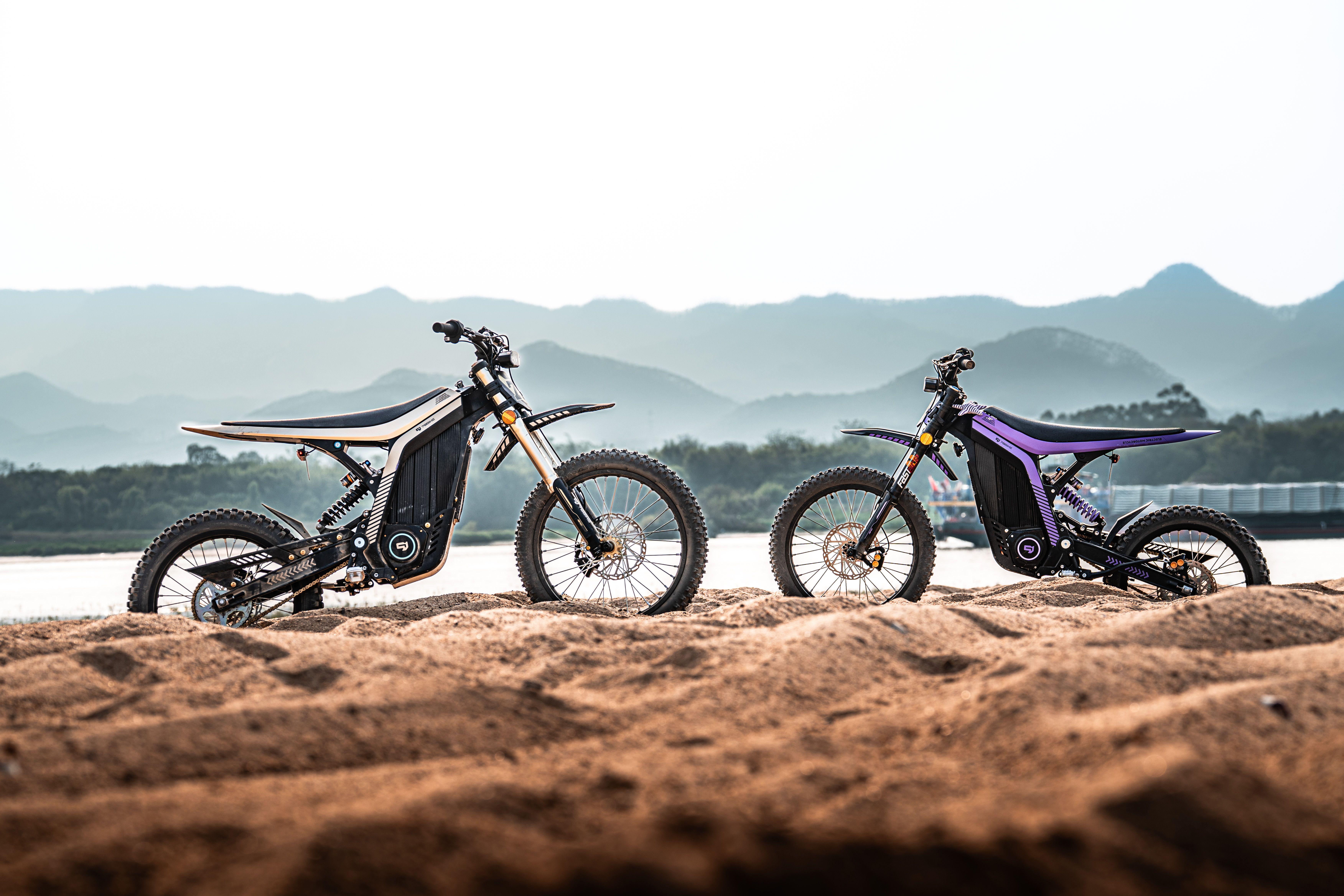 Two electric dirt bikes parked on sandy terrain against a scenic mountain backdrop during sunrise.