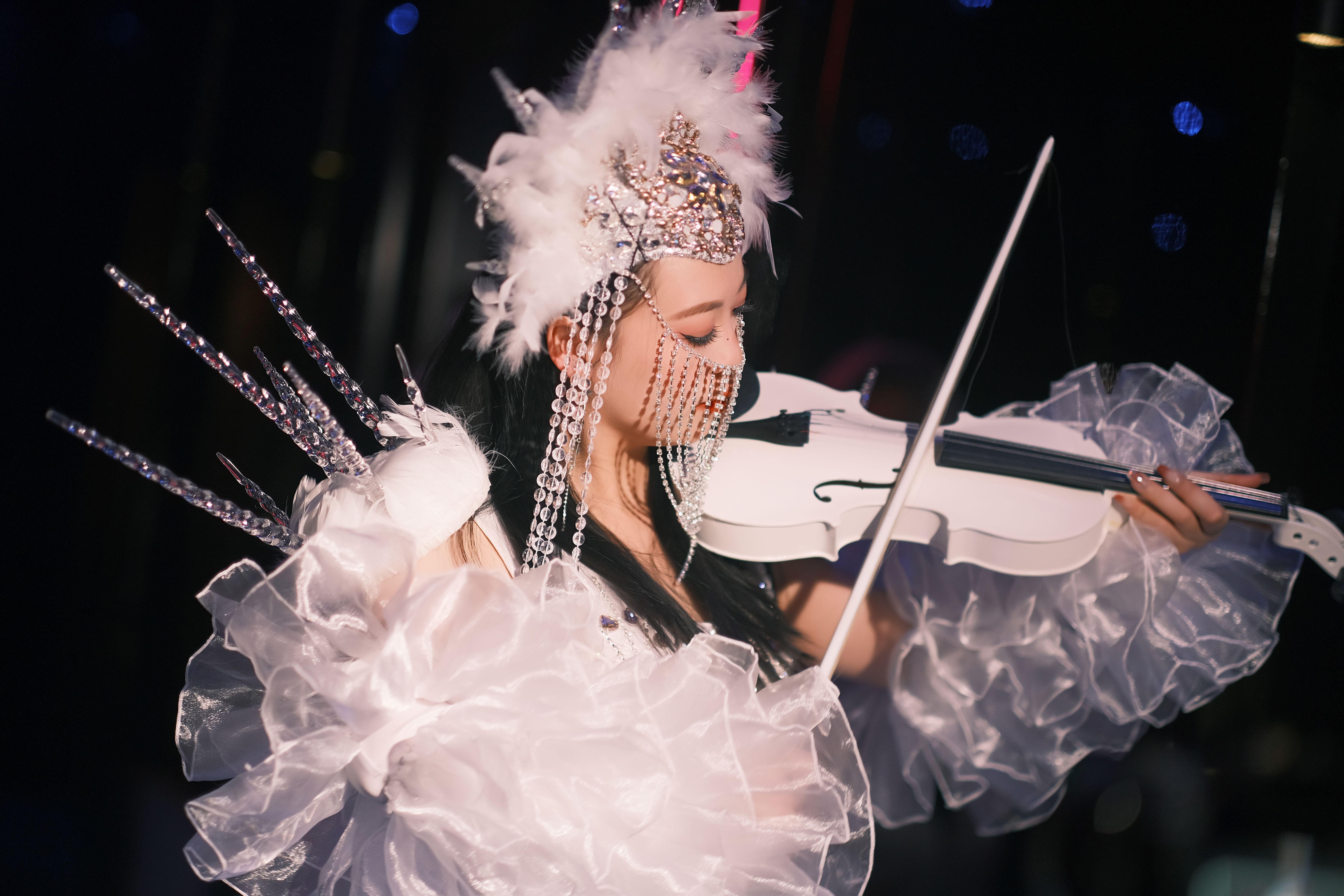 An artistic performer in elaborate costume plays a white violin under stage lights, creating a vibrant spectacle.