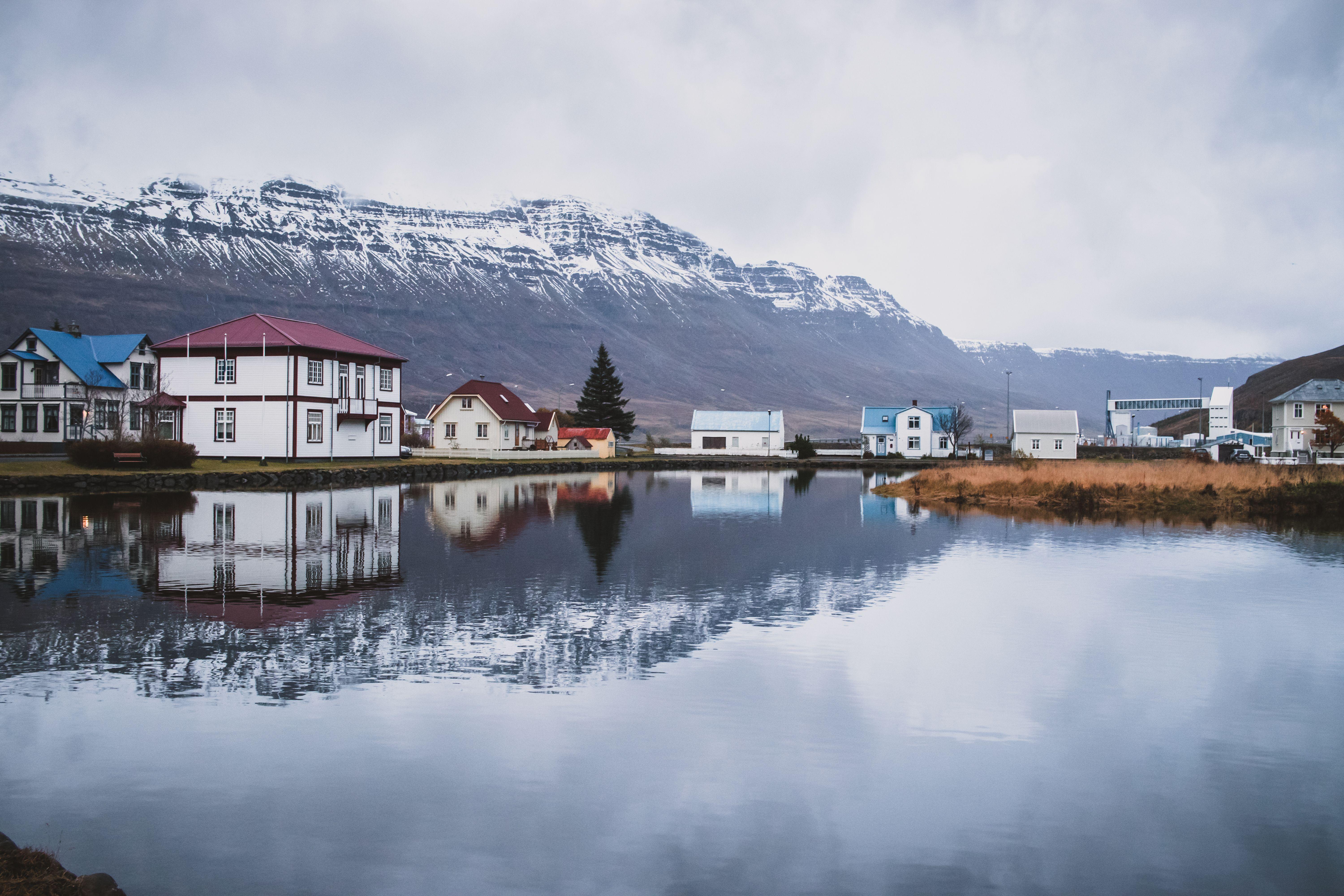 Picturesque Icelandic village with reflections on a calm lake, against a mountainous backdrop.