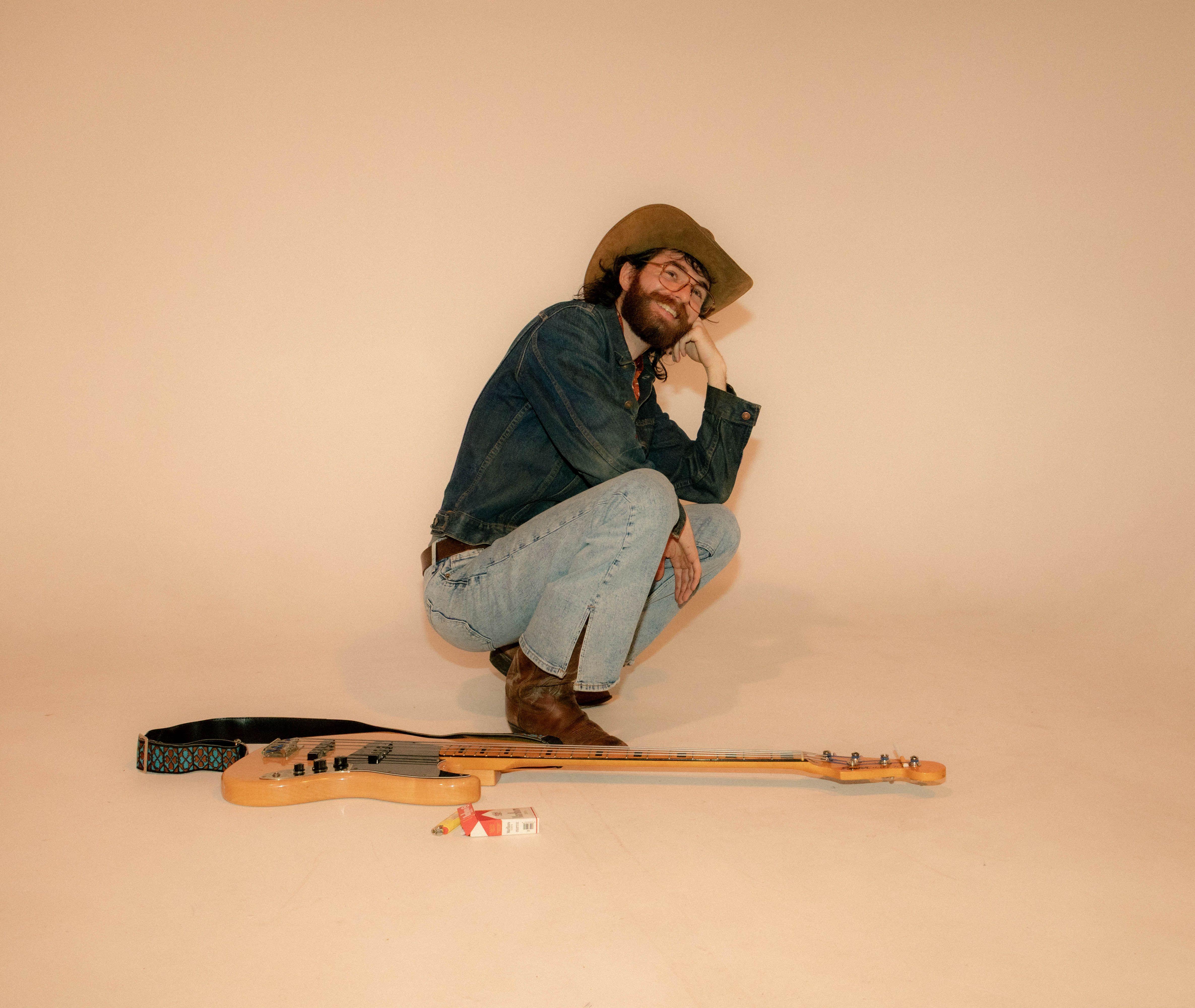 A stylish man in denim and cowboy hat crouches beside an electric guitar in a neutral-toned studio.