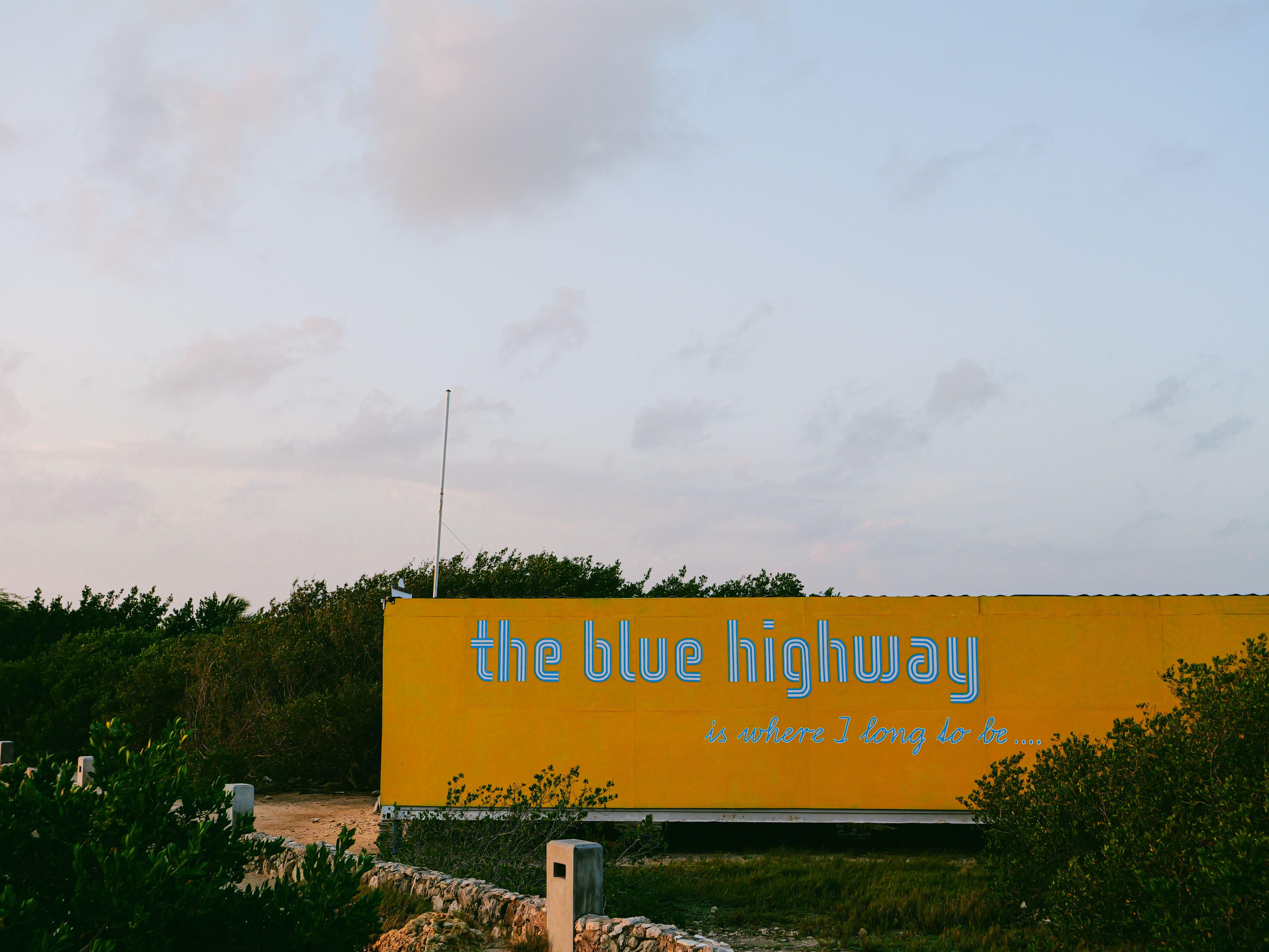 Yellow billboard with 'the blue highway' text against serene outdoor backdrop.