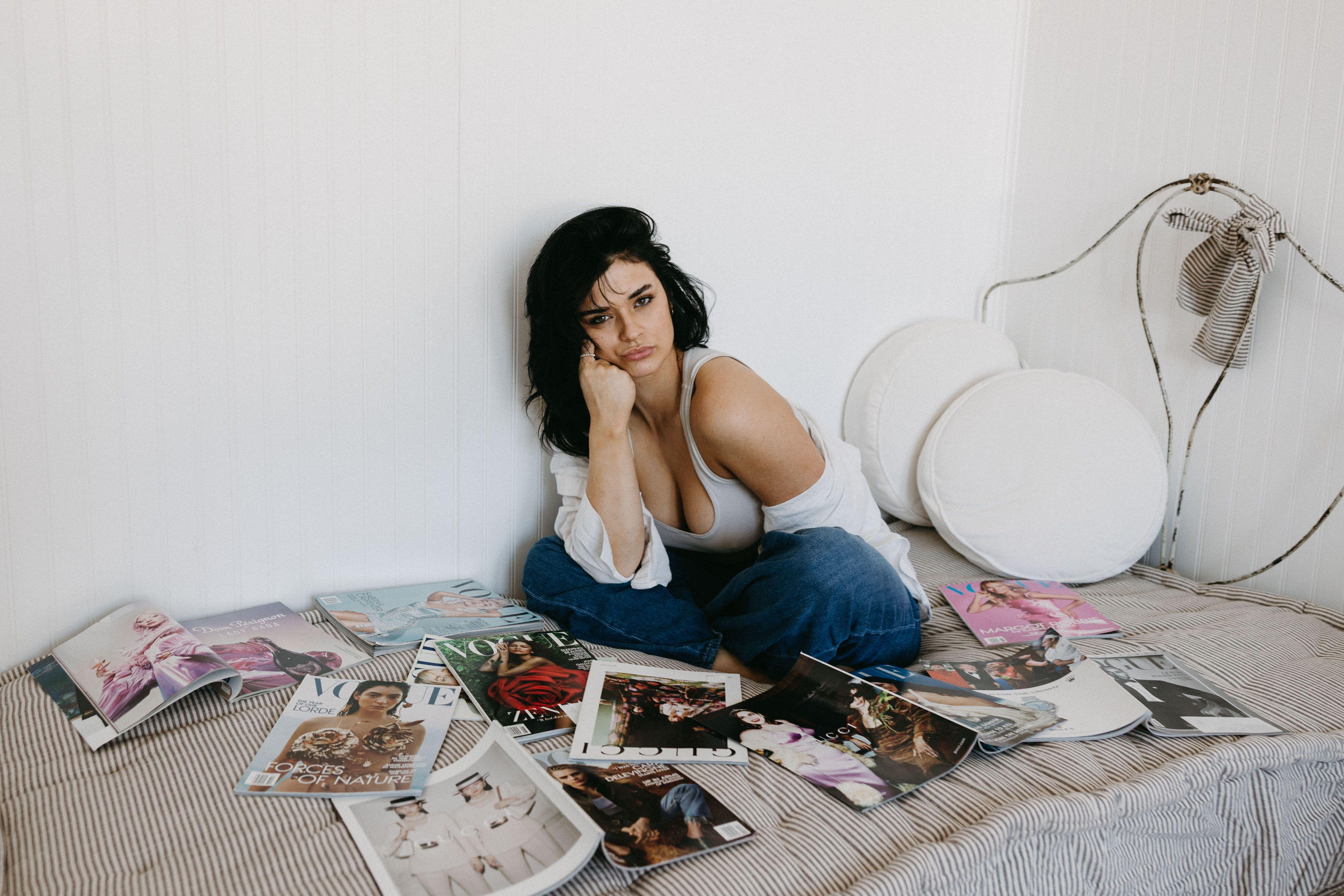 Young woman in casual attire relaxing with a collection of fashion magazines on bed.