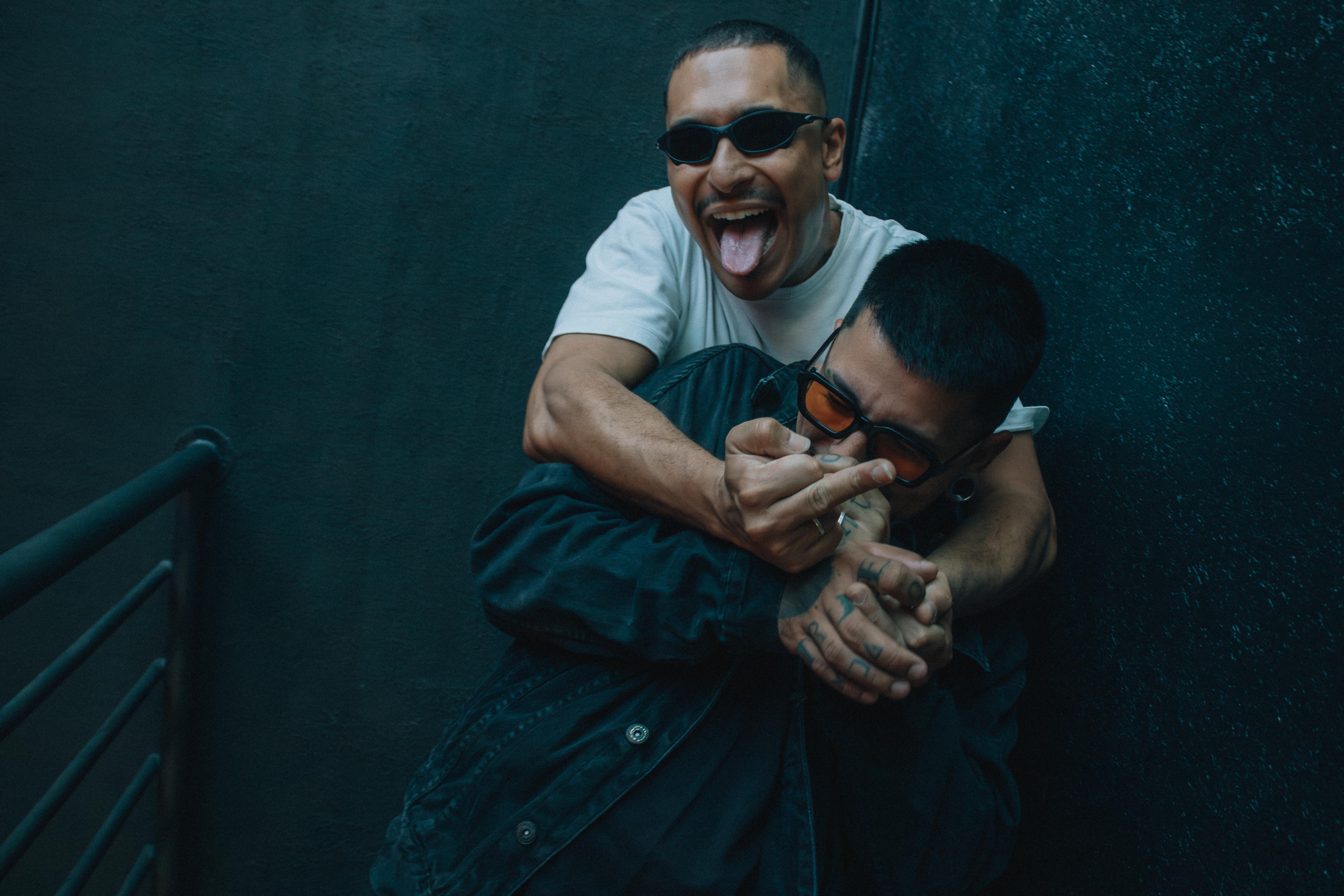 Two friends joking around indoors, wearing sunglasses and displaying playful gestures.