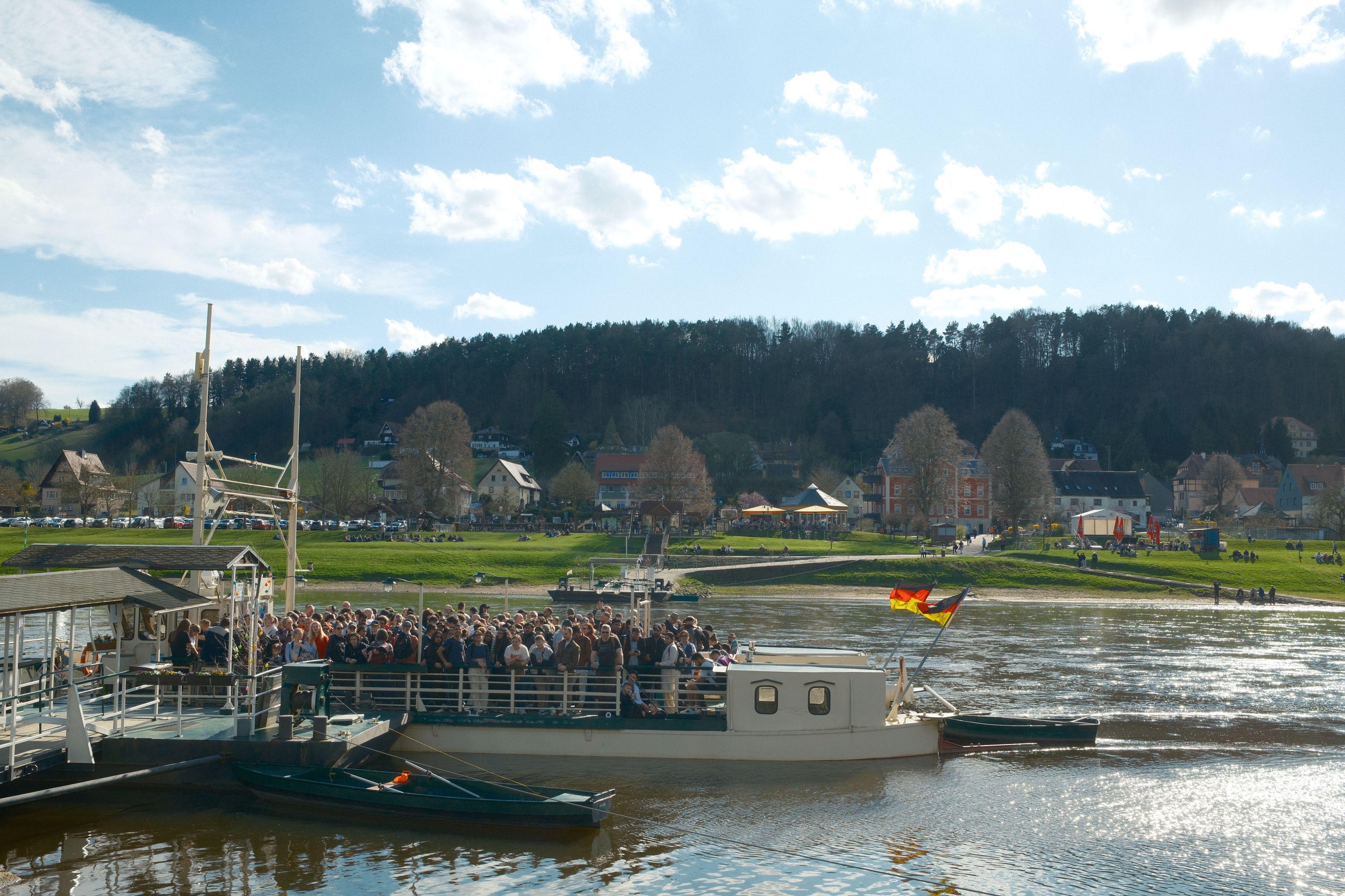 A ferry crosses a scenic river in Germany under a bright blue sky, filled with passengers.