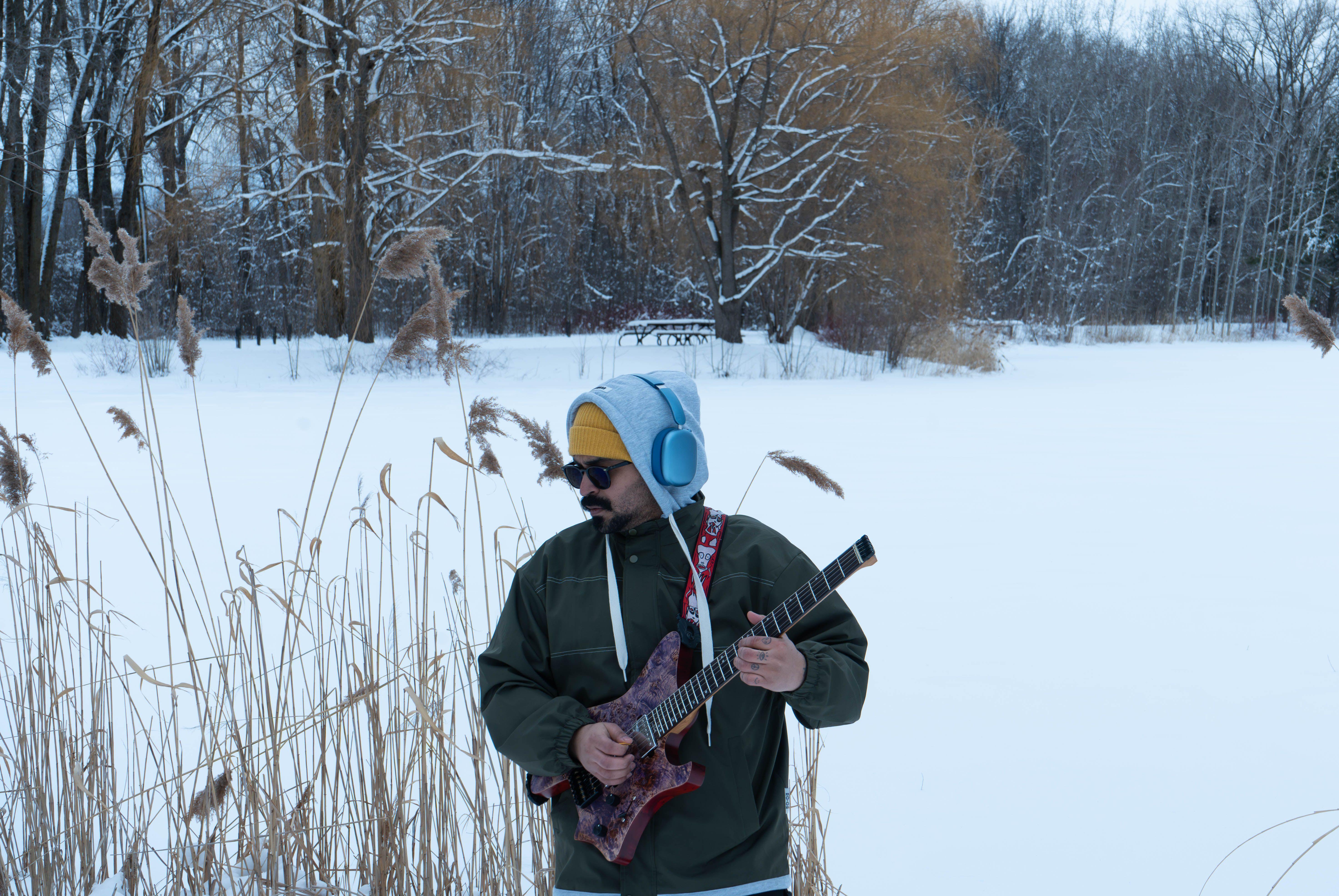 Man playing guitar in snowy park, Montreal, wearing headphones and winter clothes.