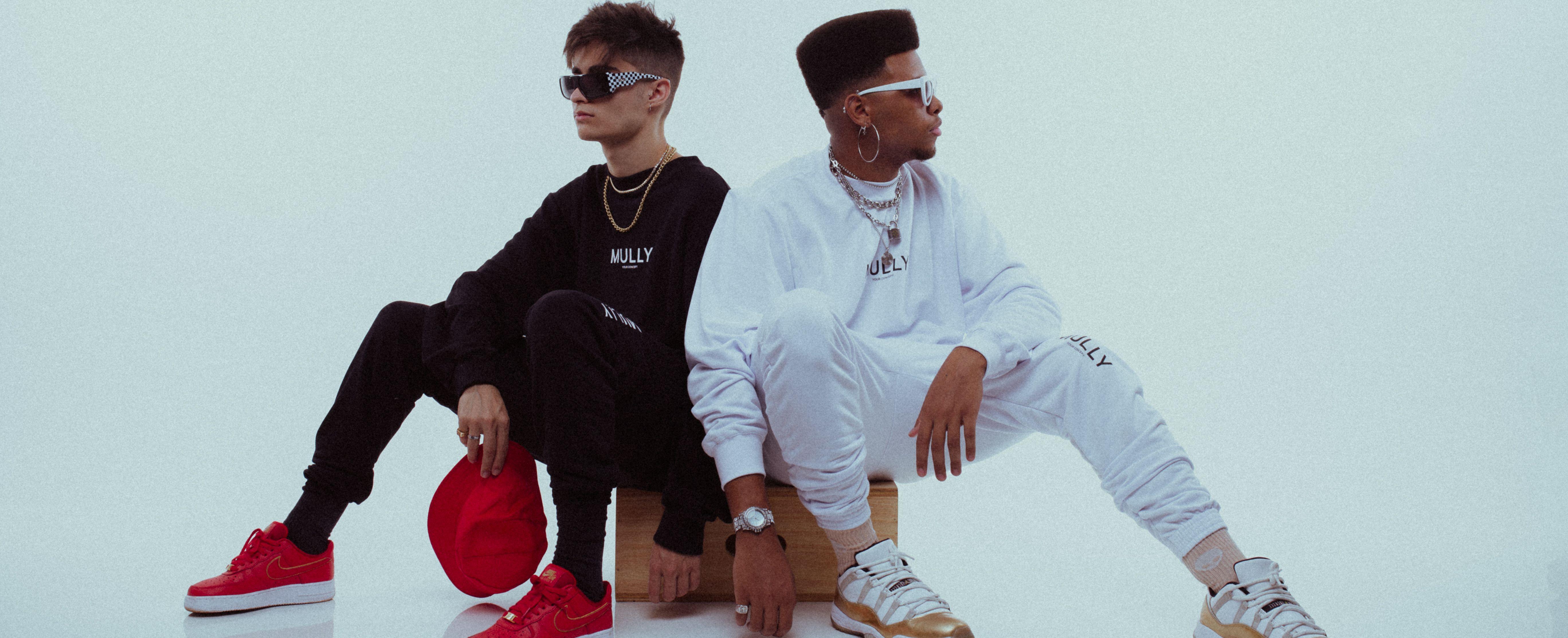 Two stylish young men sitting in a studio wearing trendy outfits and accessories, showcasing contemporary urban style.